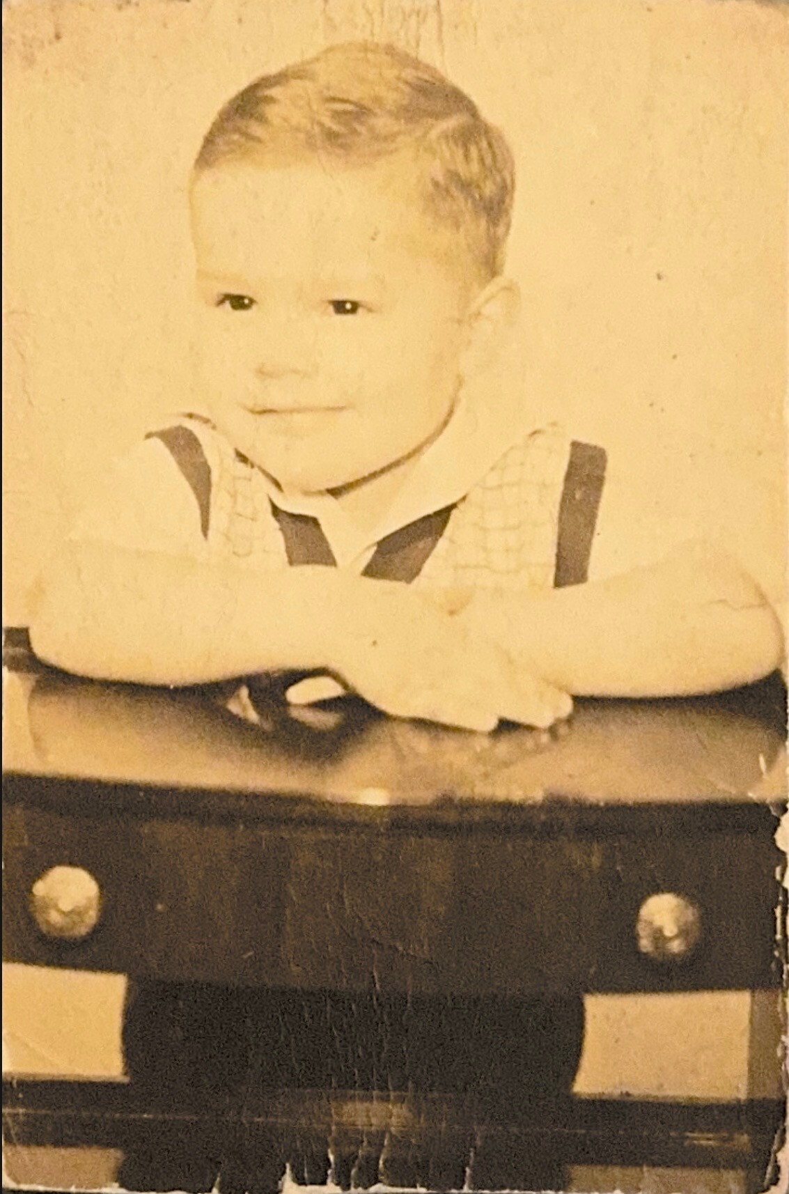 Young child with short hair resting arms on a table, wearing a light checkered shirt and dark suspenders in a sepia-toned photo.