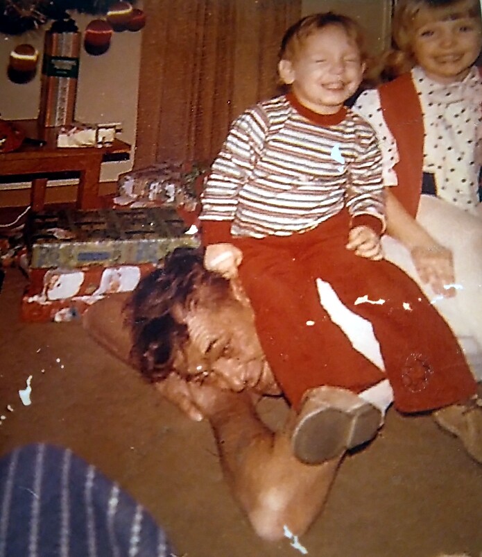Toddler in a striped shirt sits on a man's shoulders, both laughing, in a living room with wrapped gifts nearby.