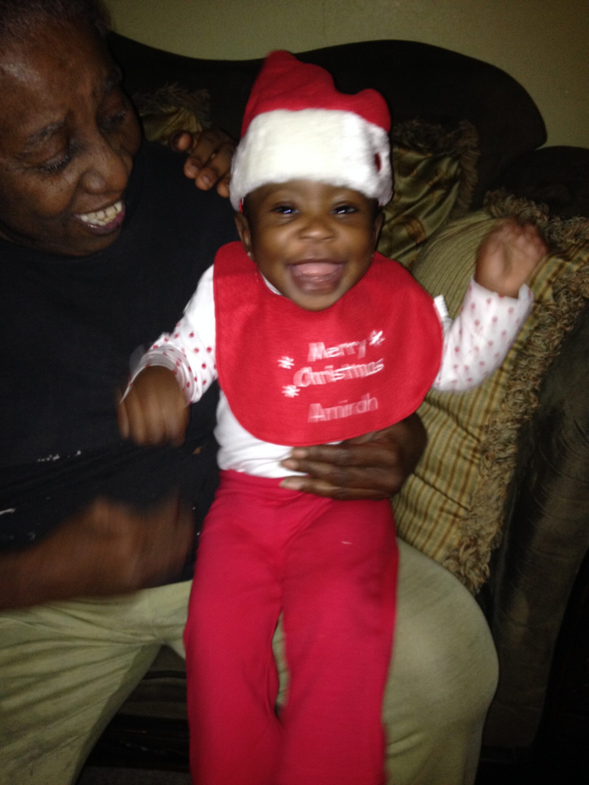 Smiling baby wearing a red Santa hat and bib, sitting on an adult's lap on a couch.