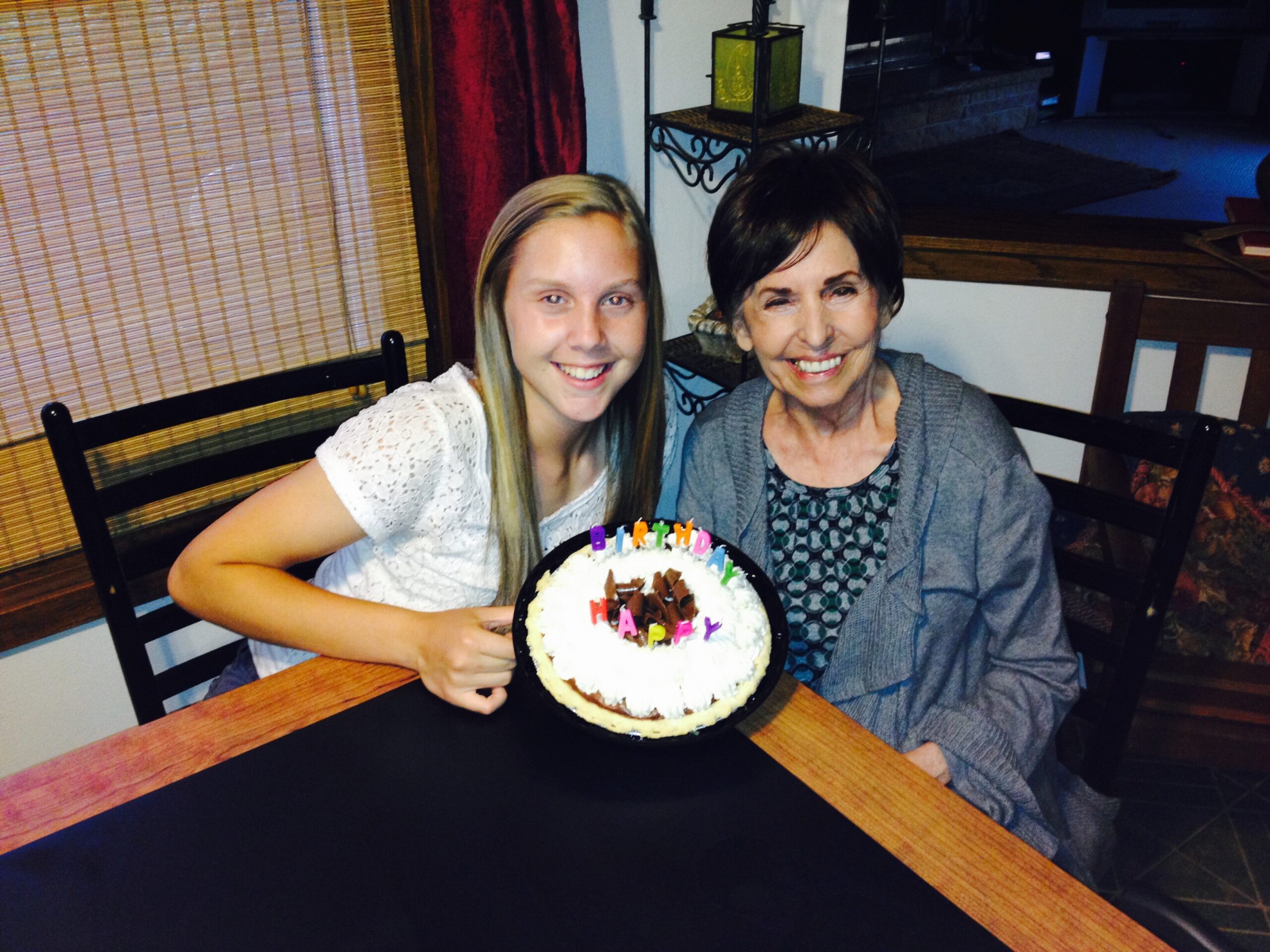 Two women smiling at a round birthday cake on a wooden table, letters spelling “HAPPY BIRTHDAY” around the cake.