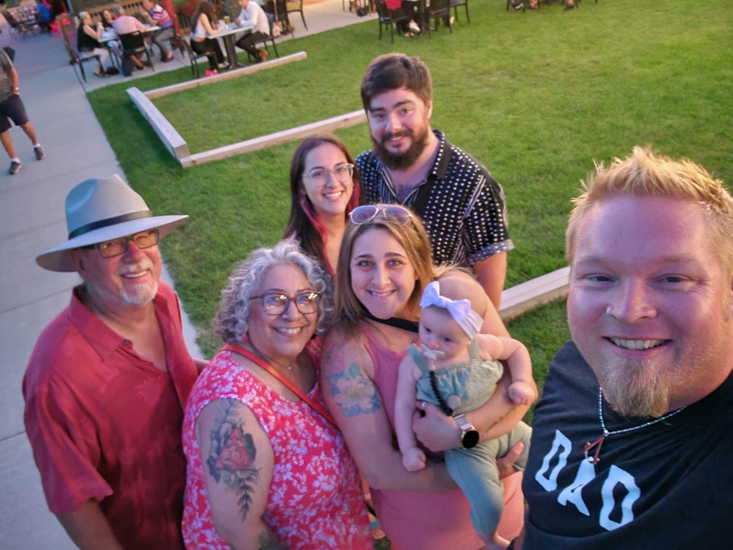 A group of six adults (plus a baby) smiling for a selfie in a park, with diners at tables in the background.
