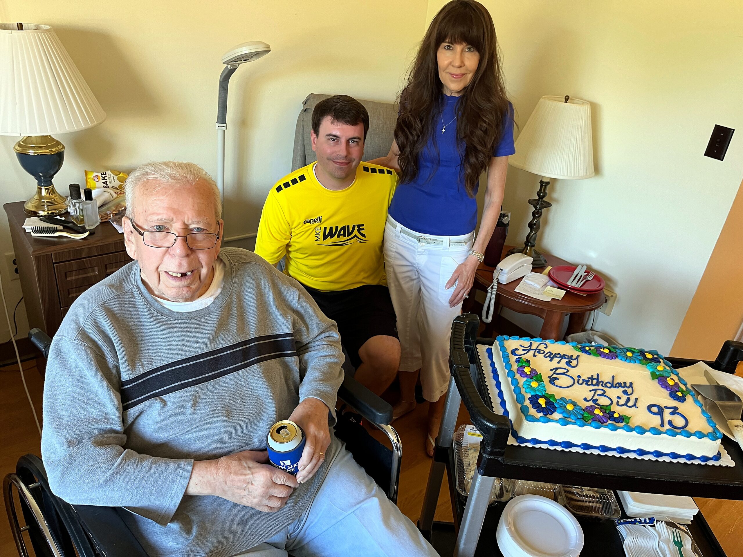 Elderly man in a wheelchair holding a drink, with a birthday cake and guests in a living room behind him.
