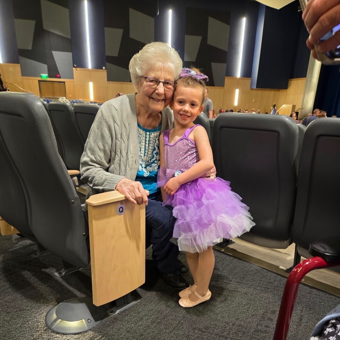 Smiling grandmother and young girl in a purple ballet tutu posing together in a theater seating area.