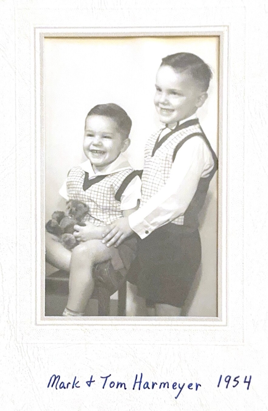 Black-and-white studio portrait of two young brothers in matching checkered vests; the younger sits holding a teddy bear, the older stands behind them with a smile. Caption reads: 'Mark & Tom Harmeyer 1954'