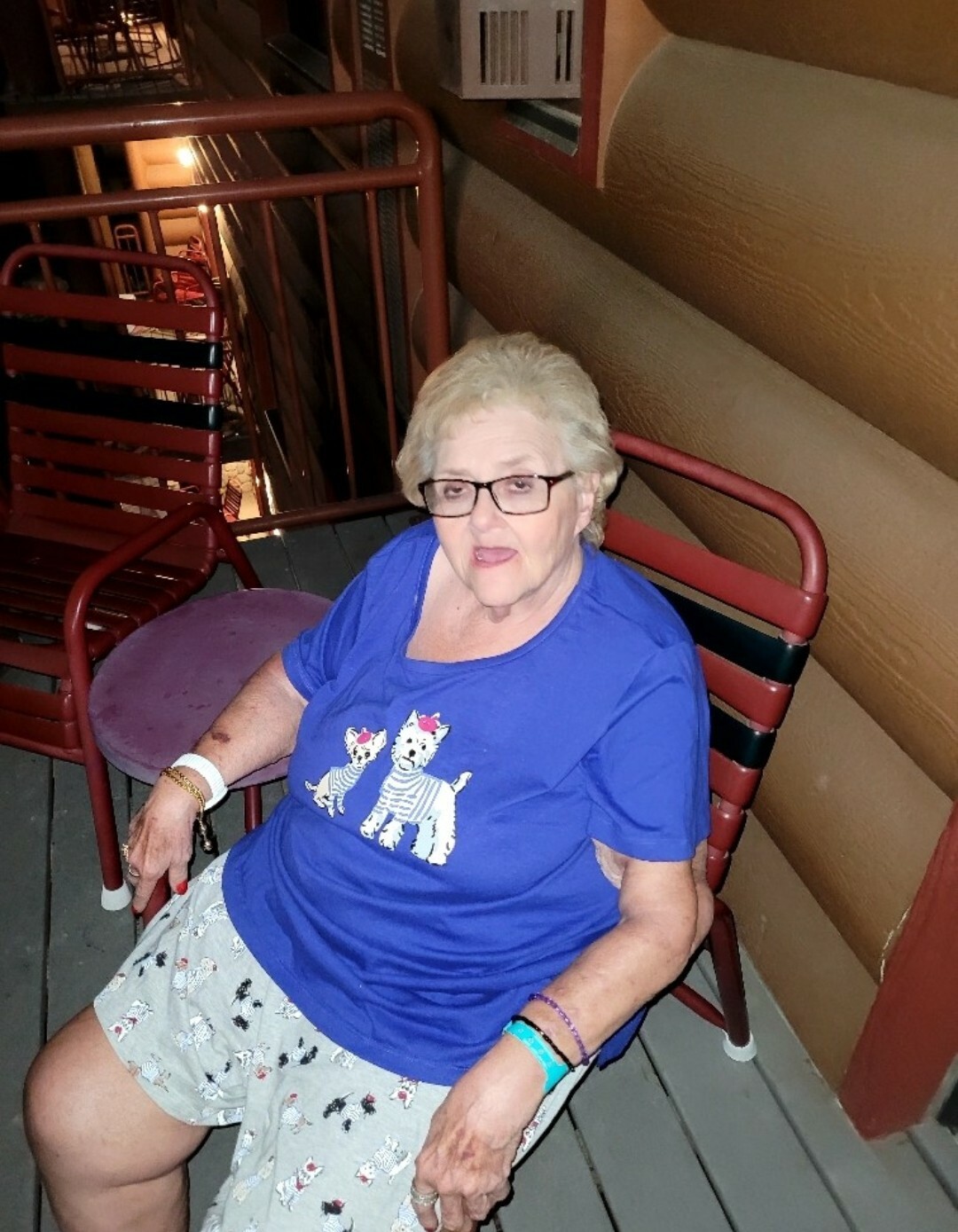 Older woman with short gray hair and glasses sits in a red metal chair on a wooden balcony, wearing a blue shirt with cat graphics and light shorts.