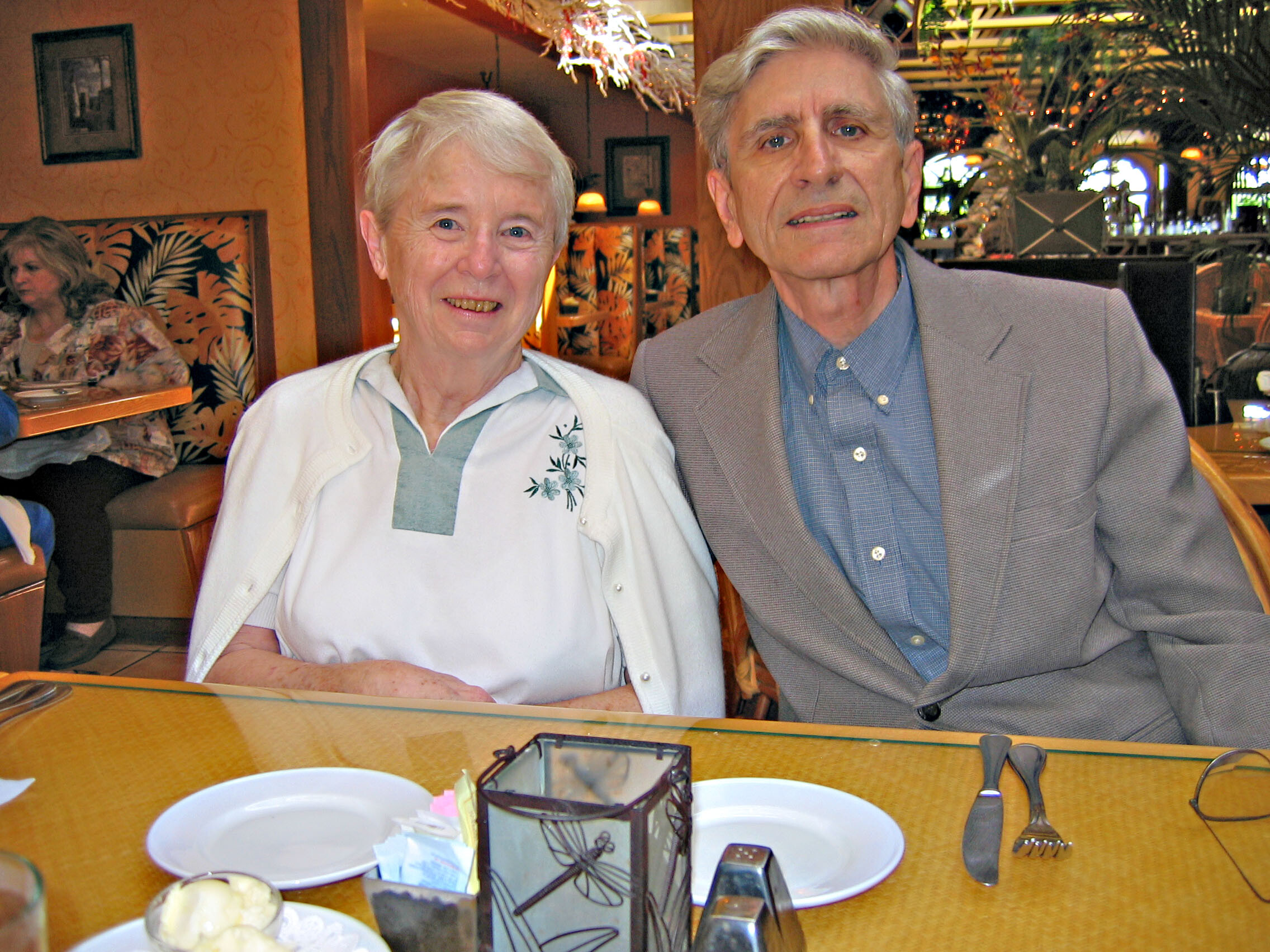 An elderly couple sitting at a restaurant table, smiling at the camera, with plates, napkin holder, and cutlery in front of them.
