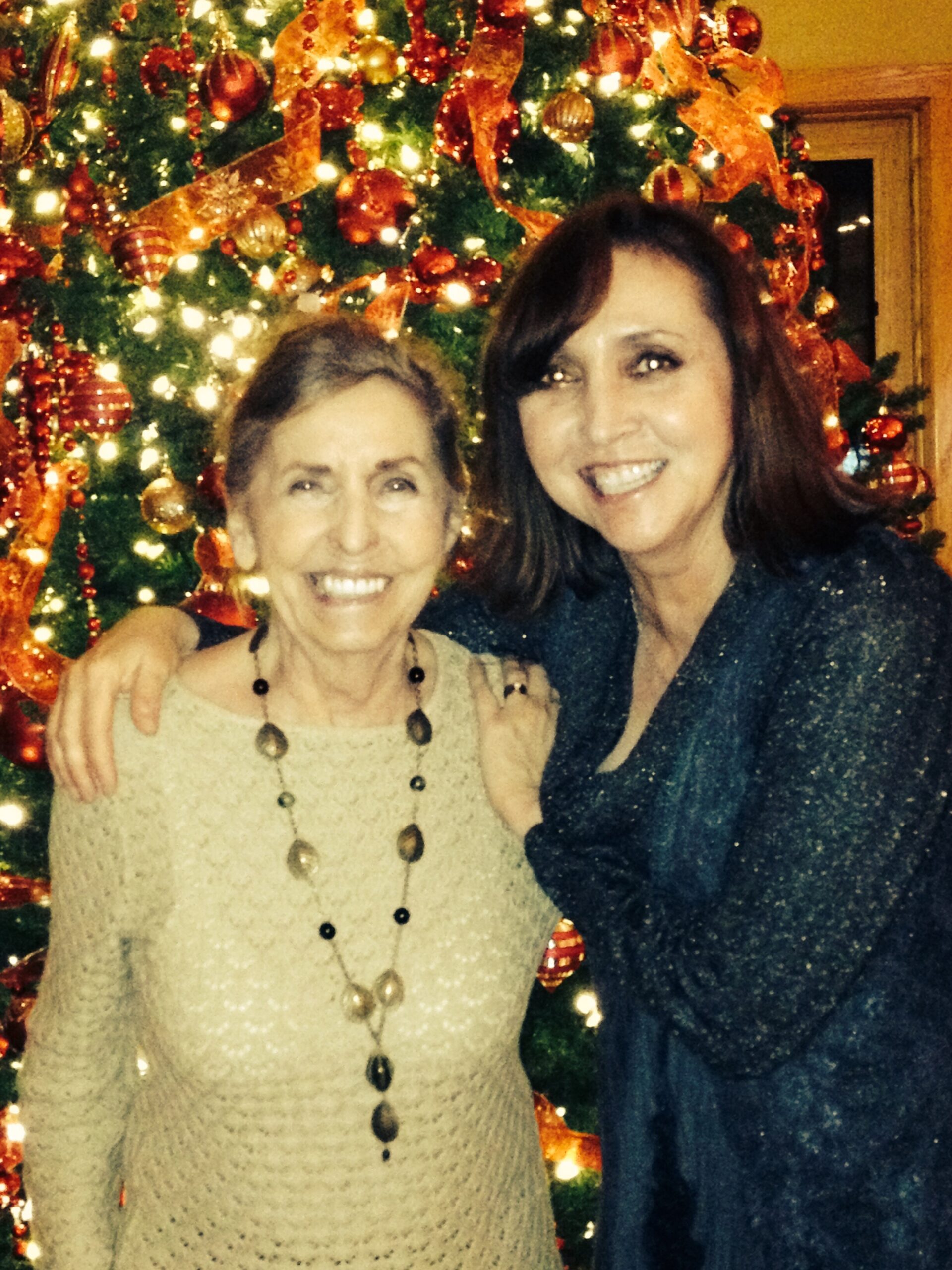 Two women smiling with arms around each other in front of a decorated, lit Christmas tree.