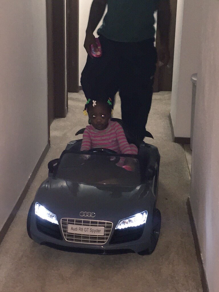 Child riding in a toy Audi R8 GT Spider in a hallway, with an adult standing behind him/her and guiding the car.