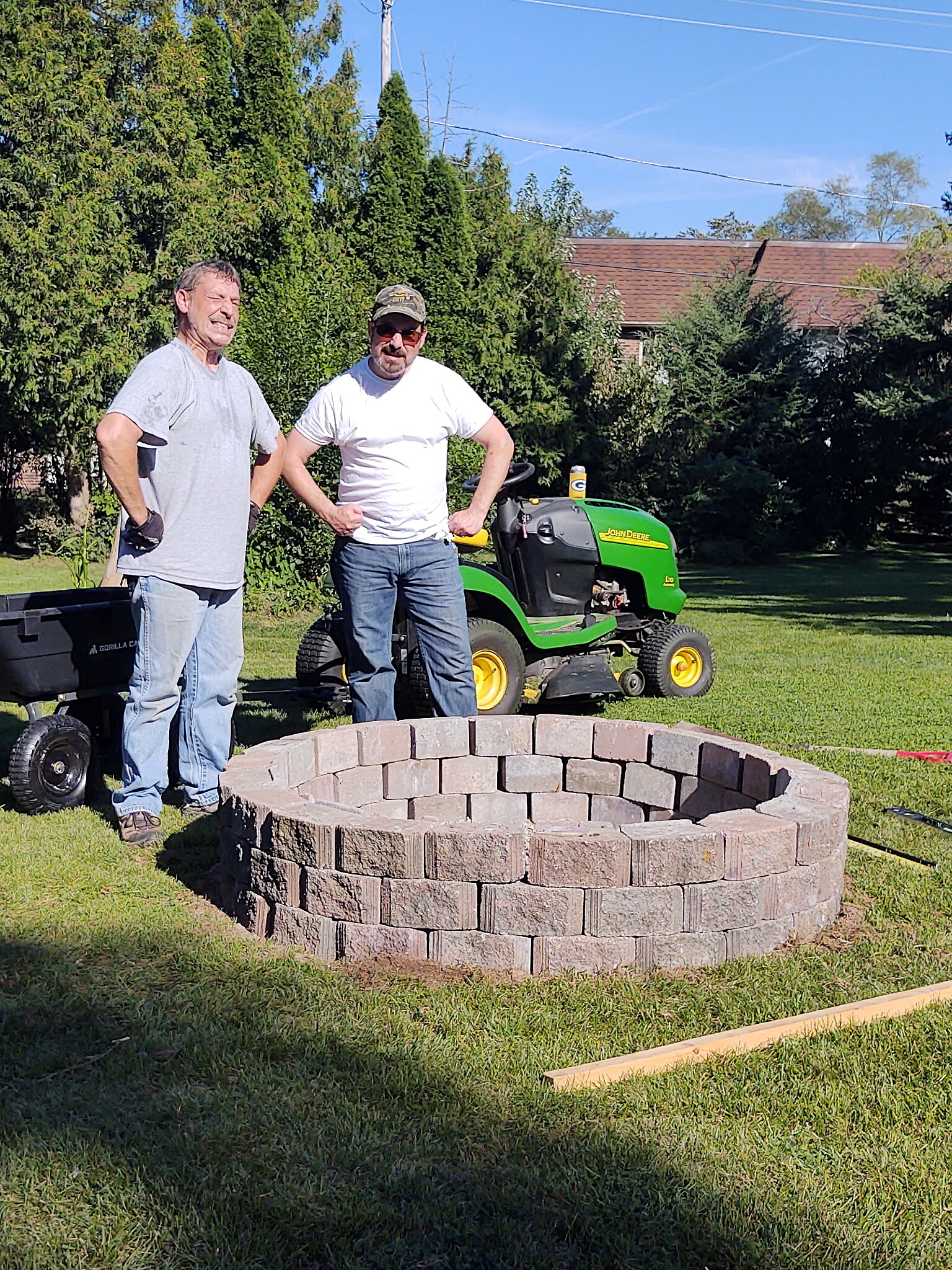 Two men stand with hands on hips beside a circular stone fire pit in a sunny backyard, a green John Deere lawn tractor behind them.