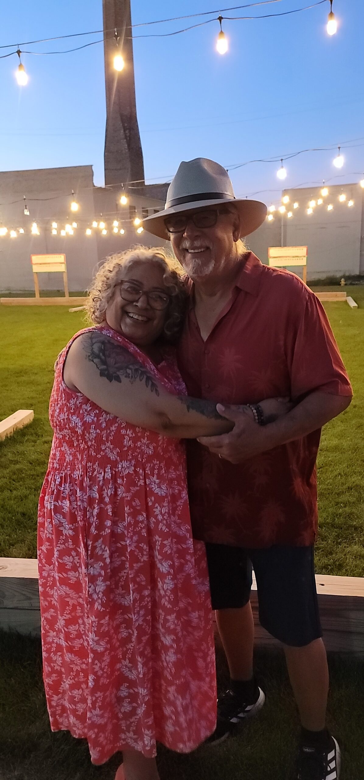 Smiling couple embraces at an outdoor evening event beneath string lights at dusk.