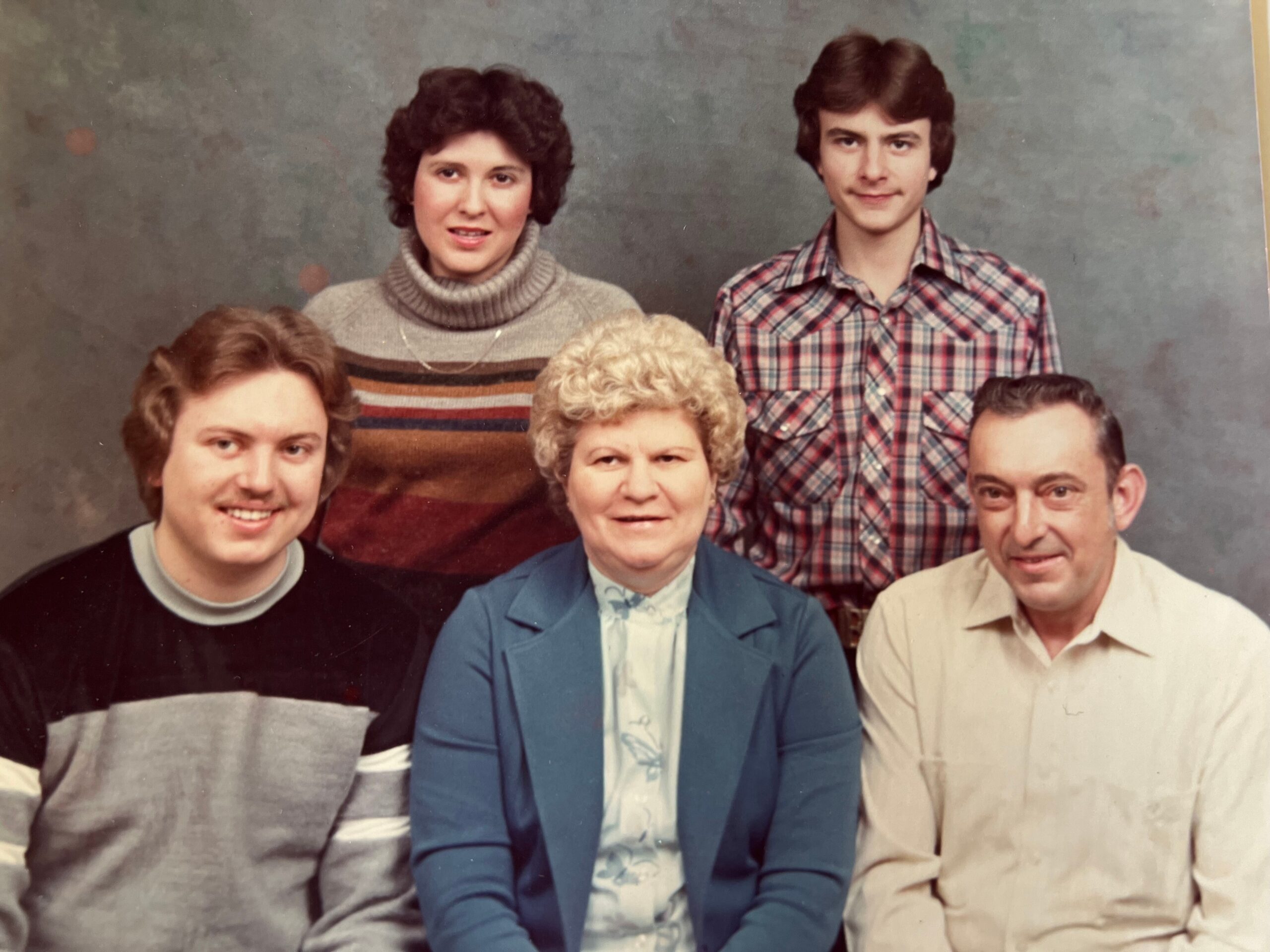 Family group portrait in a studio, five people posed in two rows, wearing 1980s clothing and smiling.