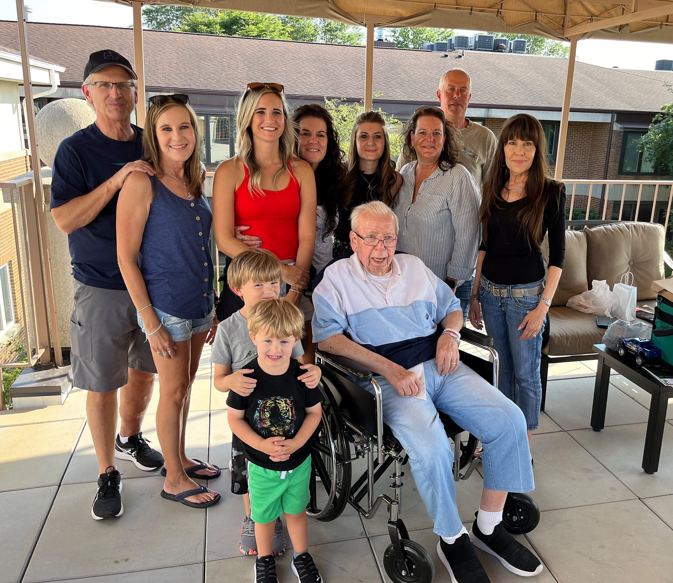 Smiling group photo on a sunny balcony: several adults standing behind two children and an elderly man in a wheelchair in front. two people wear casual clothes and sunglasses while others pose around them.