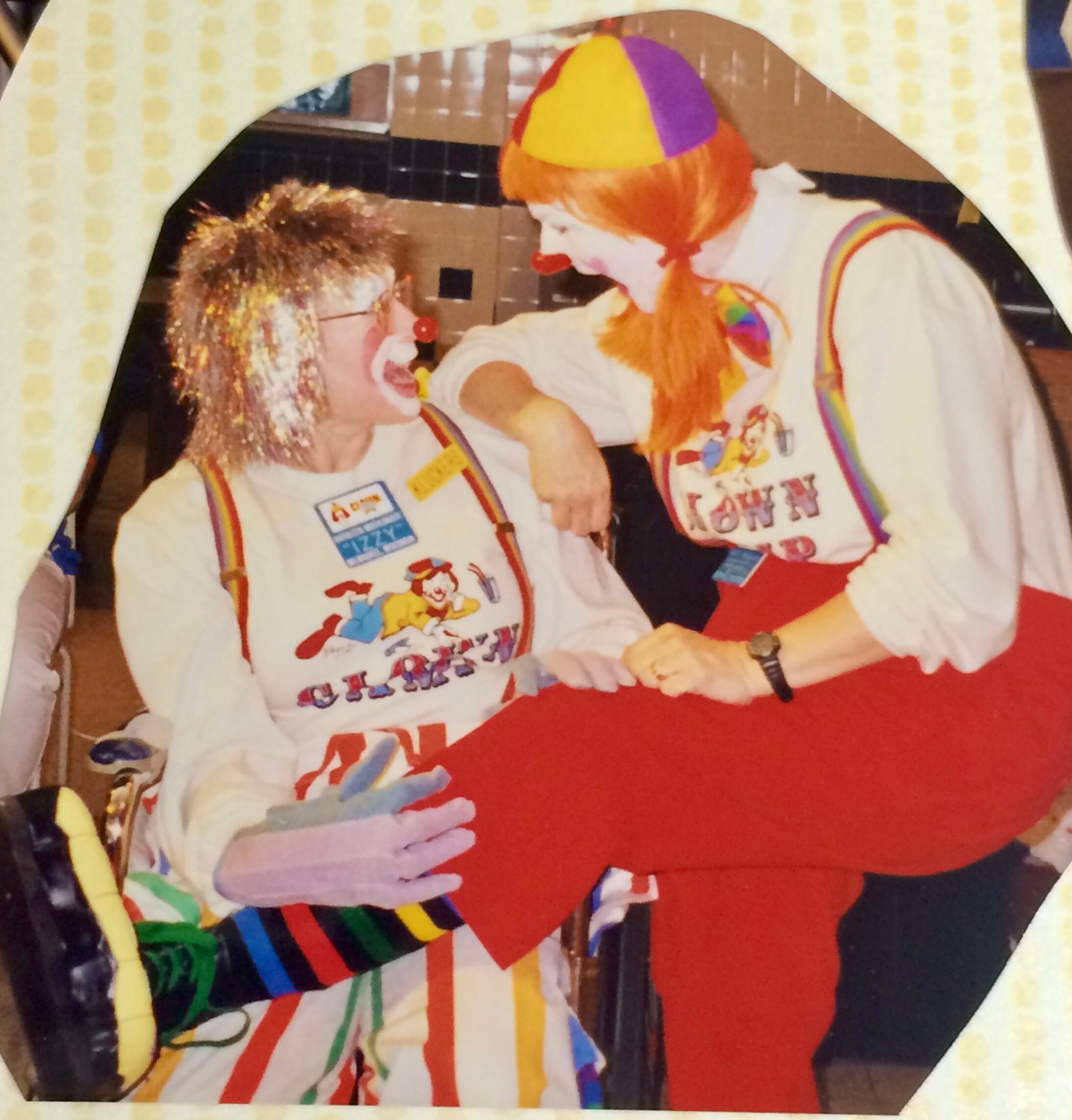 Two costumed clowns share a moment, wearing white shirts with cartoon designs, rainbow suspenders, and bright hats at a festive event backlit by colorful decor.