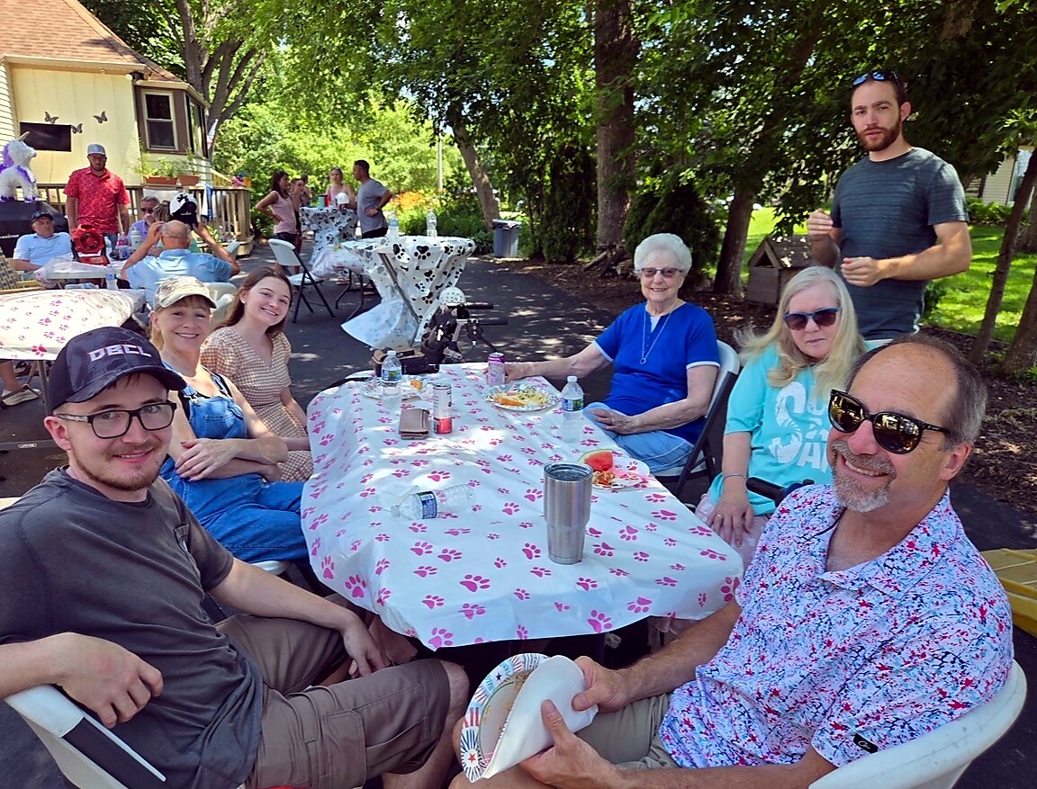 Group of friends and family seated around a backyard picnic table with a pink paw-print tablecloth, enjoying an outdoor gathering on a sunny day.