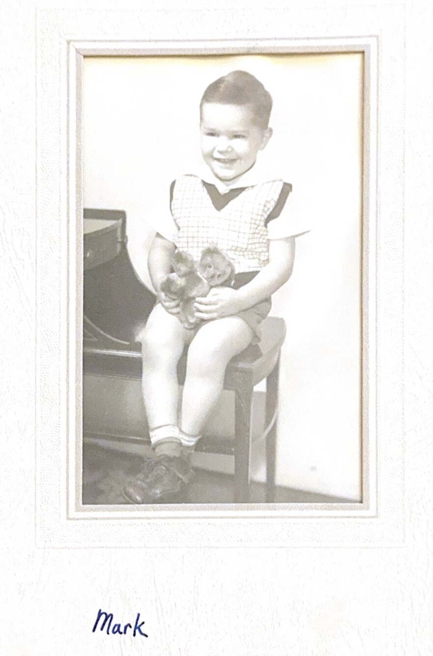 Young boy sits on a chair, smiling, holding two teddy bears; wearing a checkered vest and shorts.