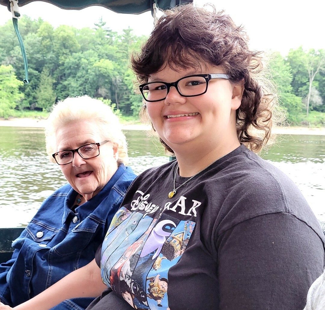 Two smiling women sit on a boat by a lake with a forested shoreline in the background; younger woman wears glasses and a black graphic t‑shirt, older woman wears a denim jacket and glasses.