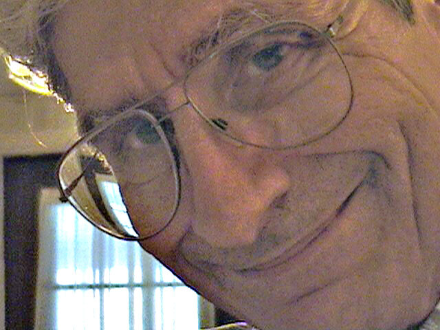 Close-up portrait of a man with round glasses and a warm smile, tilting his head toward the camera.