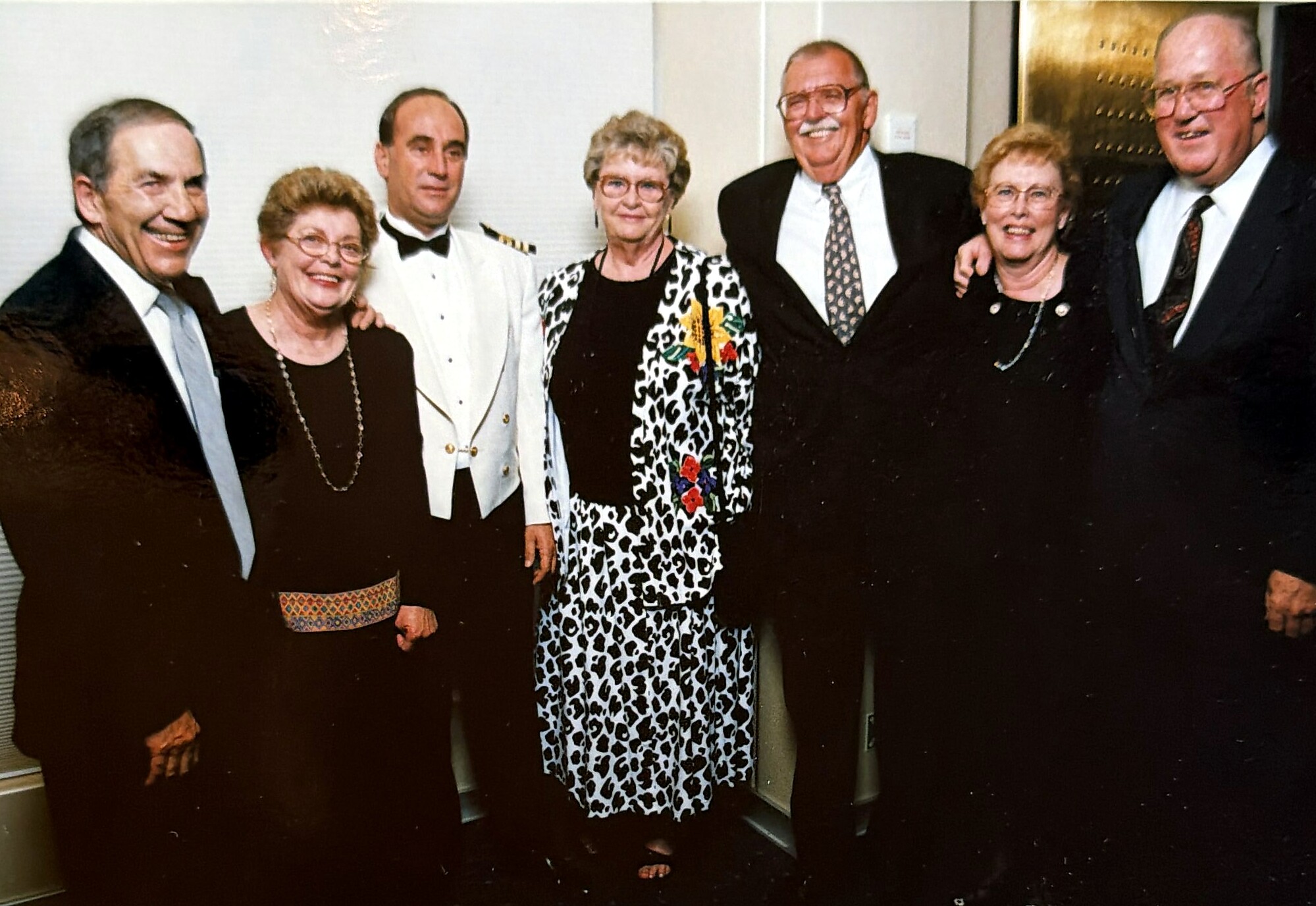 Group of seven older adults in formal attire posing for a photo at a social event; central woman wears a leopard print suit with a corsage, flanked by men in tuxedos and women in black dresses.