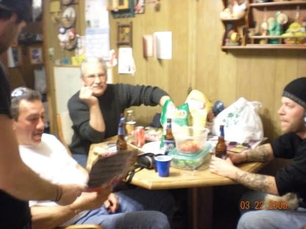 Four men sit around a cluttered wooden table indoors, drinking beer and chatting.