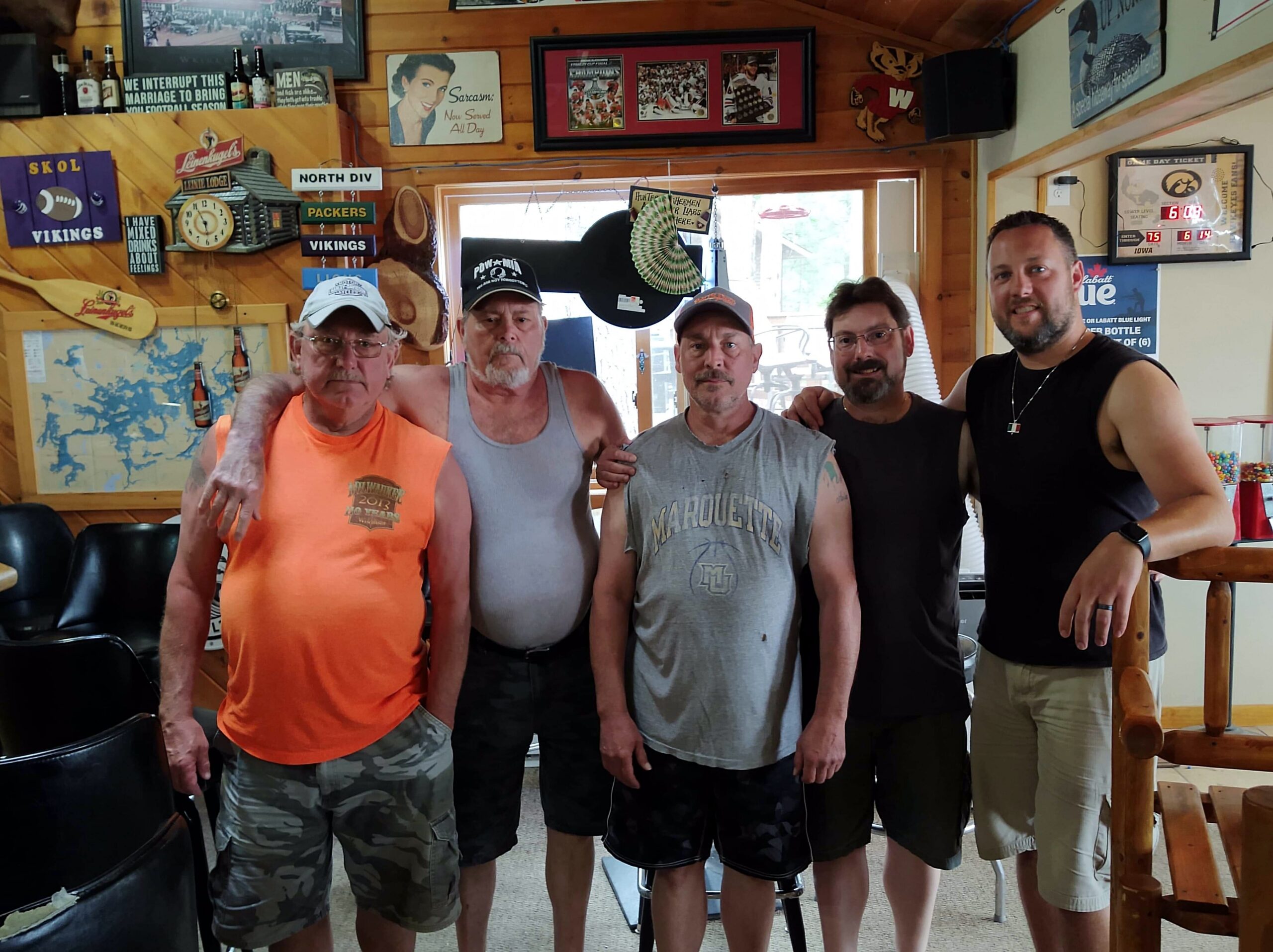 Five men stand shoulder to shoulder in a wood-paneled bar, posing for a photo with sports memorabilia on the walls behind them.