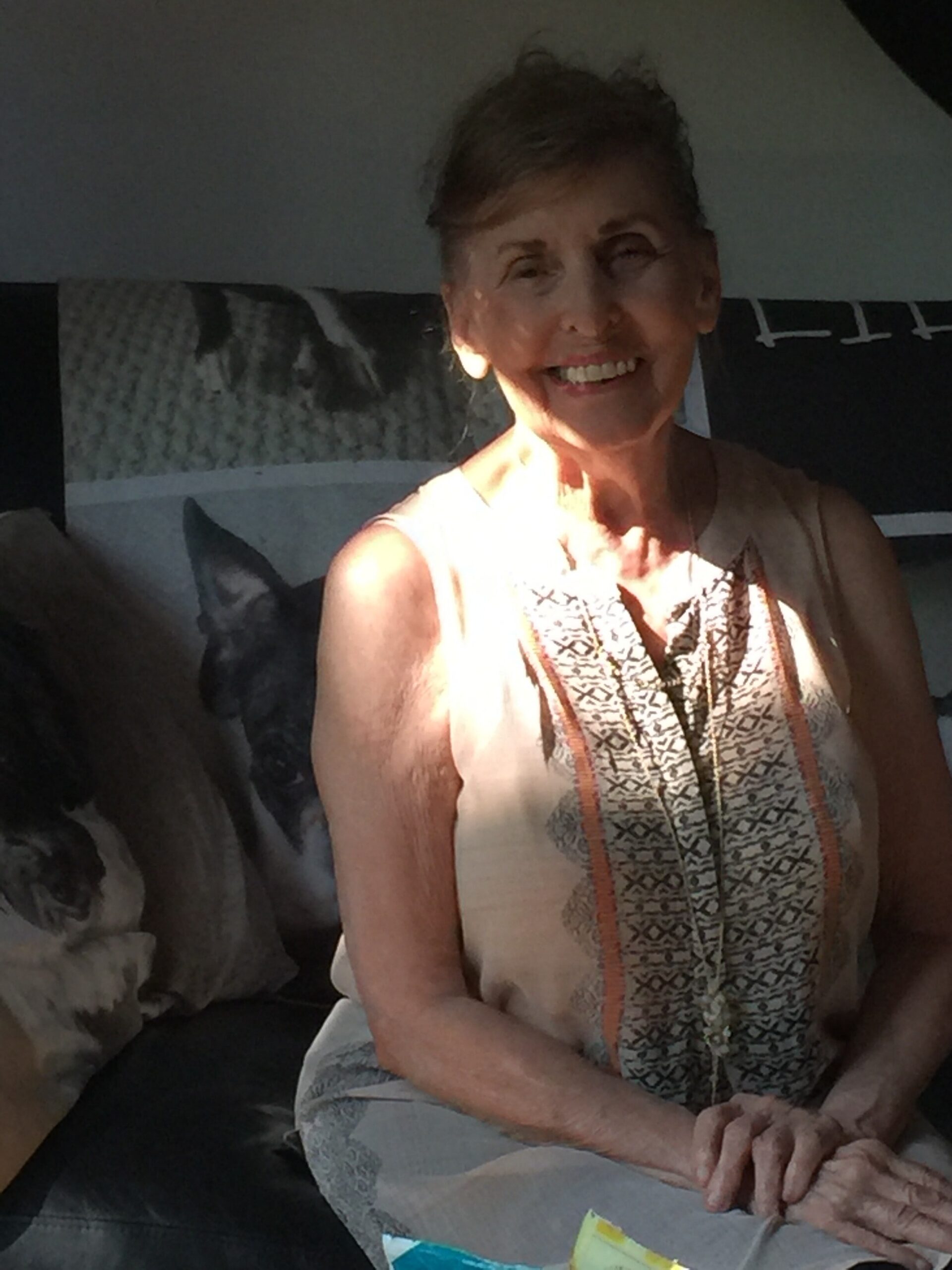 Smiling elderly woman with short hair, wearing a patterned sleeveless top, sitting indoors with hands folded in her lap.