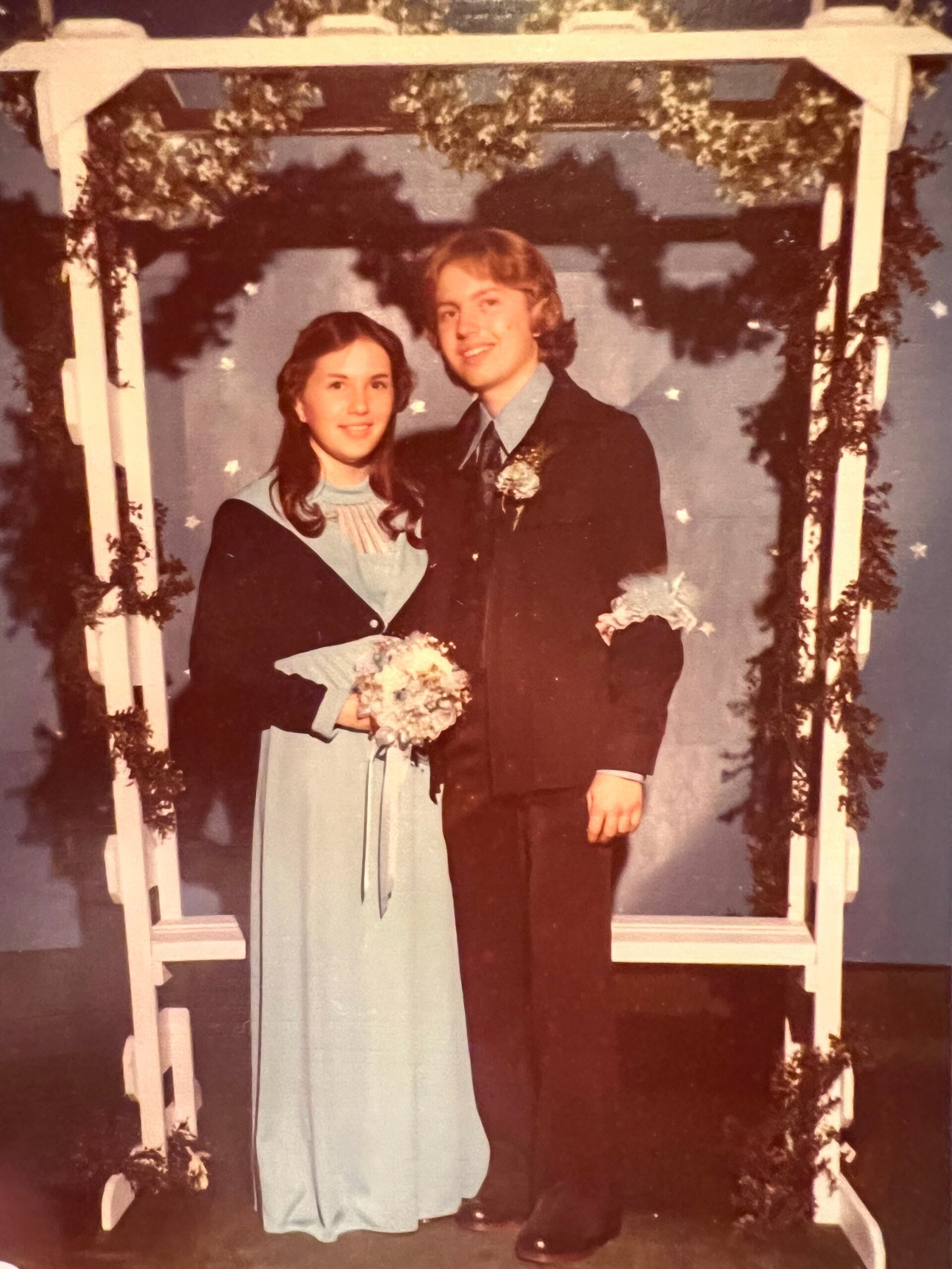 Couple in formal attire posing under a white, greenery-decorated archway.