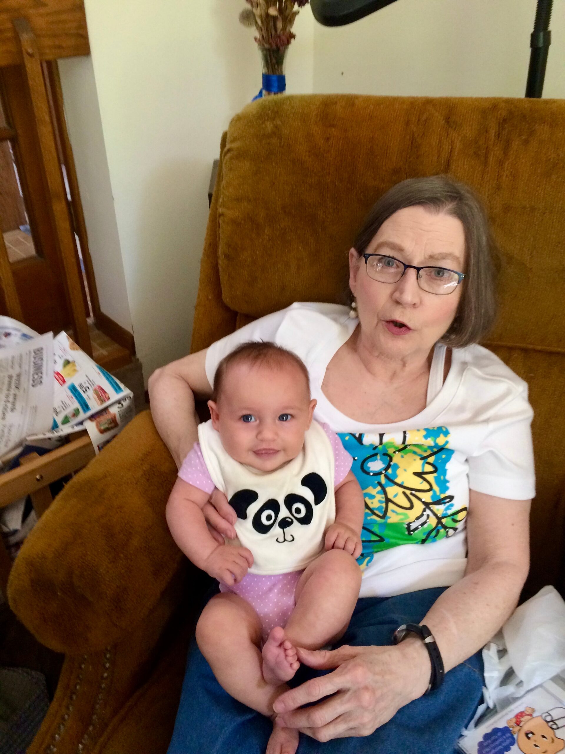 Older woman with short gray hair and glasses holds a smiling baby in a panda outfit on a brown recliner.