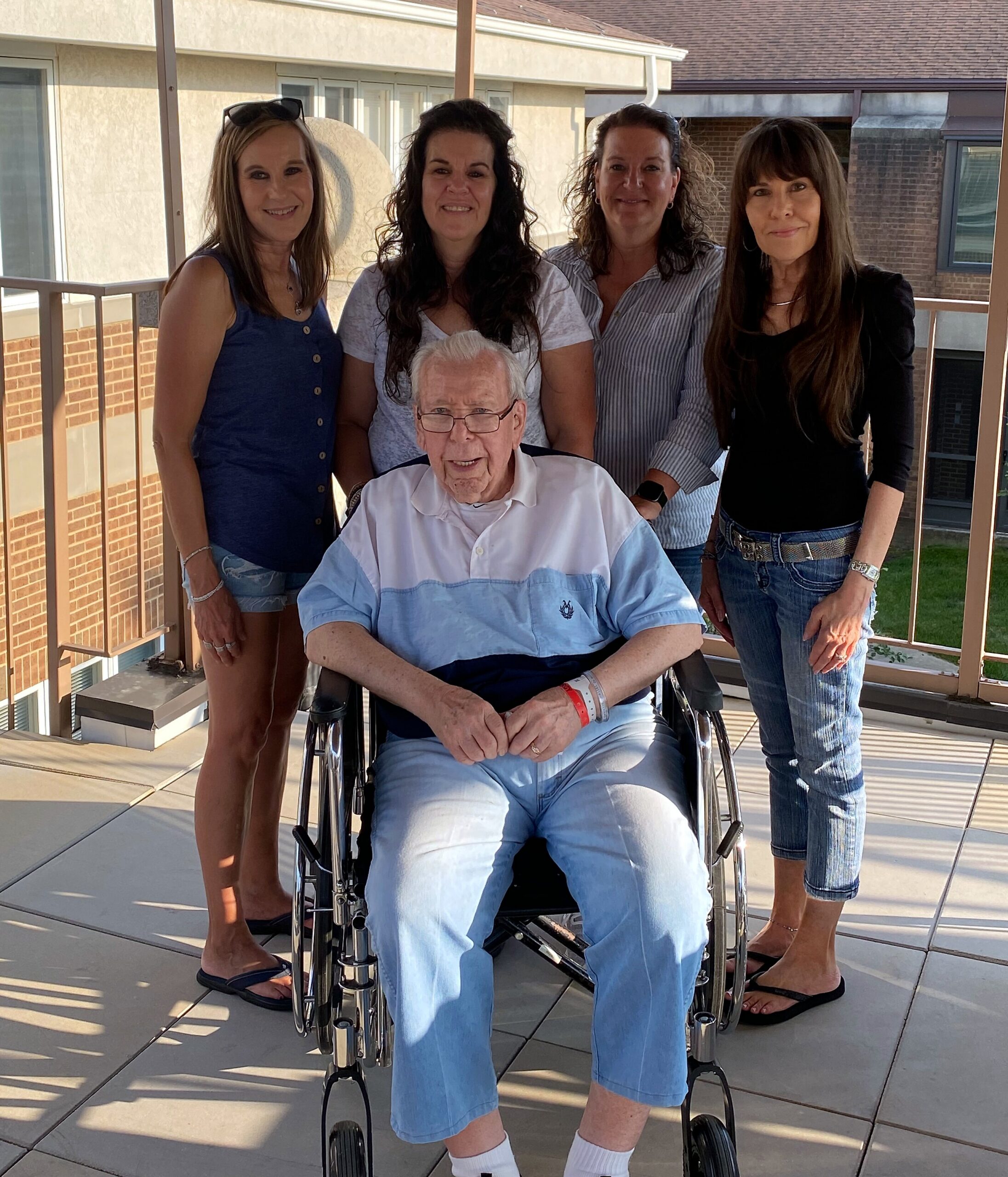 Older man in a wheelchair flanked by four smiling women on a sunny balcony.