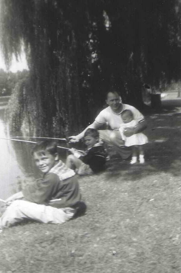 Black-and-white photo of a father with two children and a baby fishing along a lakeshore under a large tree; they watch the water together.