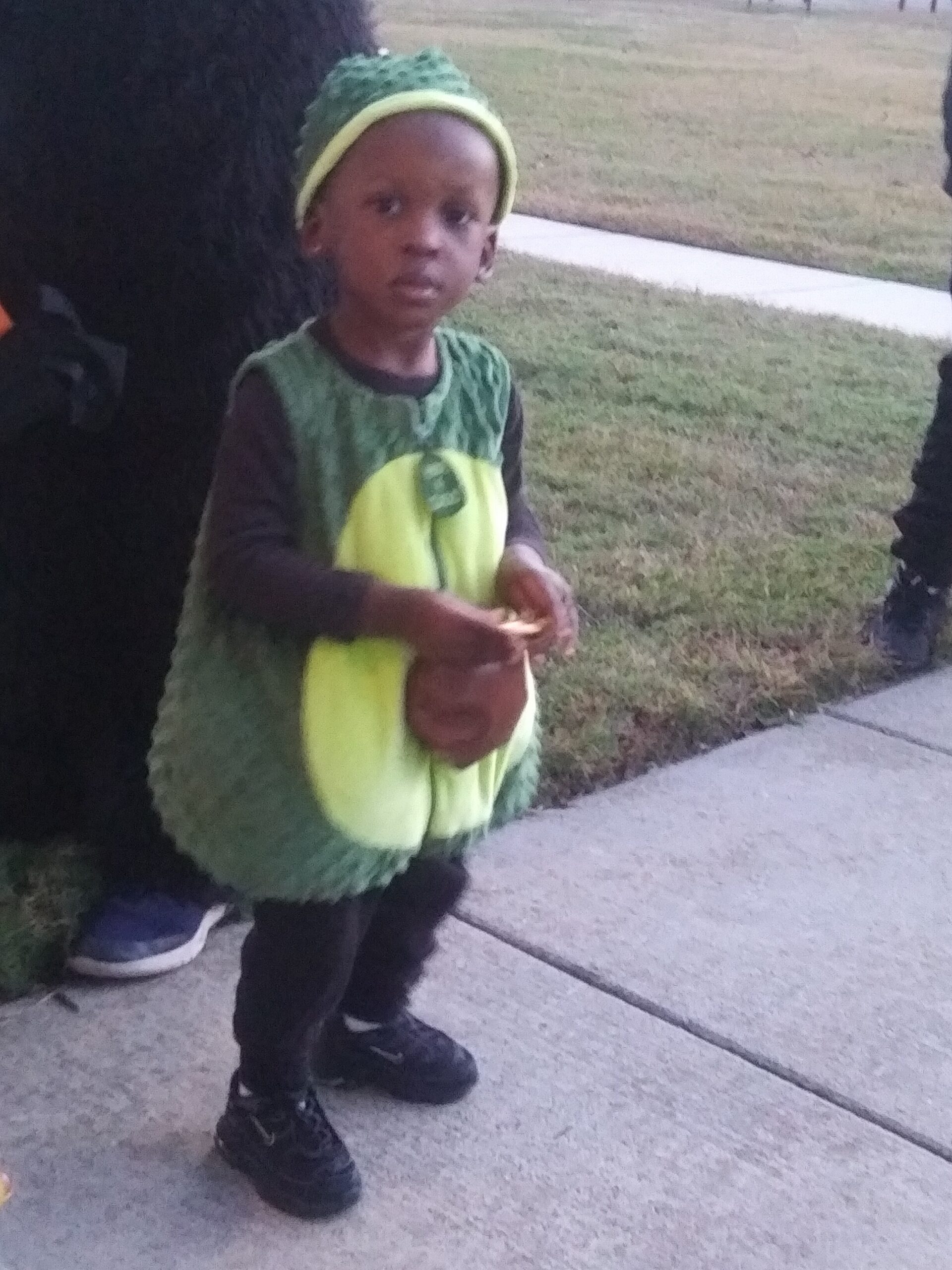 Young child in a green dinosaur costume with a yellow belly, standing on a sidewalk outdoors, wearing a matching headband and sneakers.