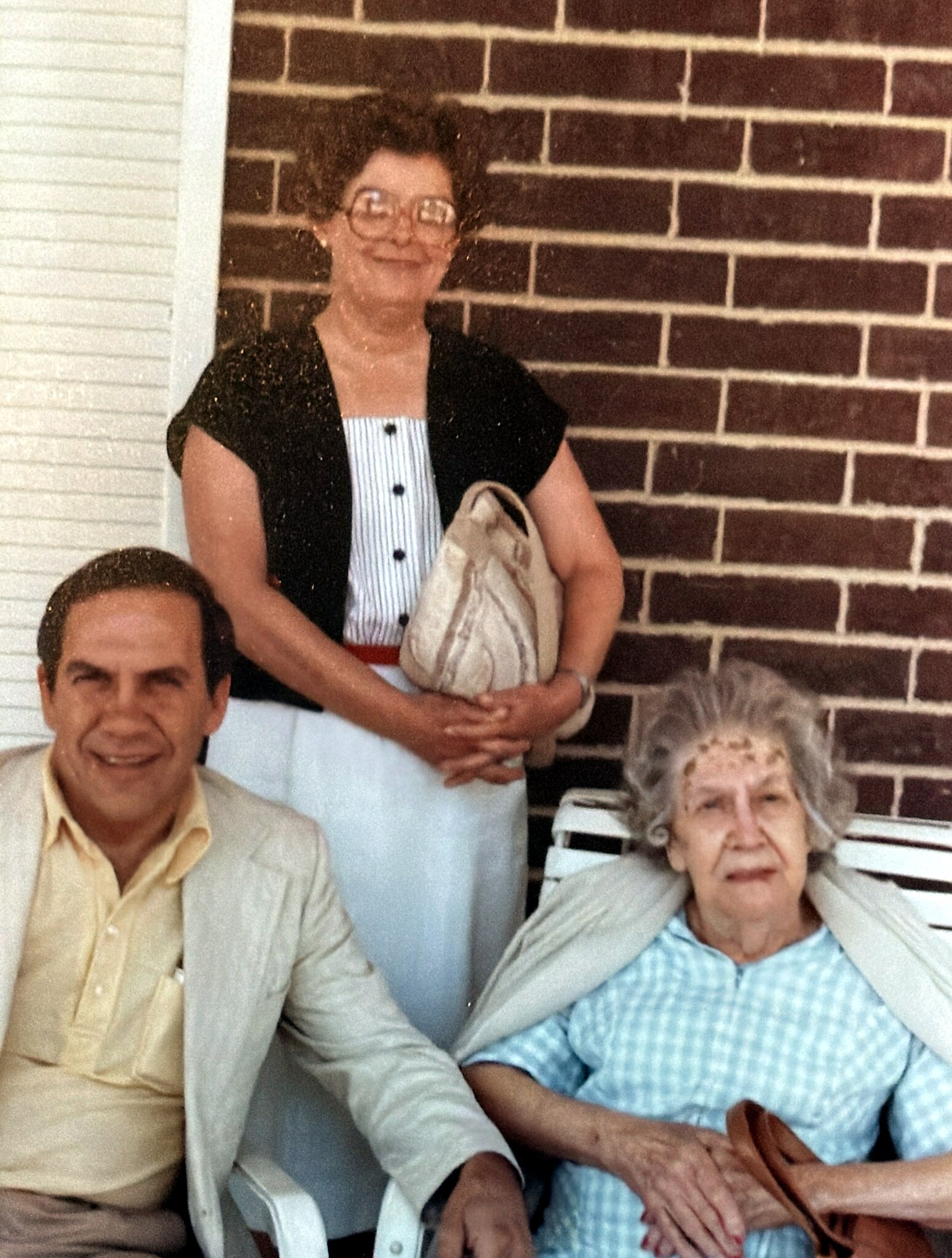 Three older adults posing on a porch with a brick wall: a man seated left, a standing woman in the center holding a bag, and an elderly woman seated right.