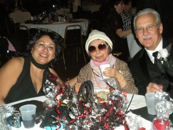 Three people sit at a banquet table smiling: a woman in black on the left, an elderly woman with a white hat and sunglasses in the center, and a man in a suit on the right, with festive decorations on the table.