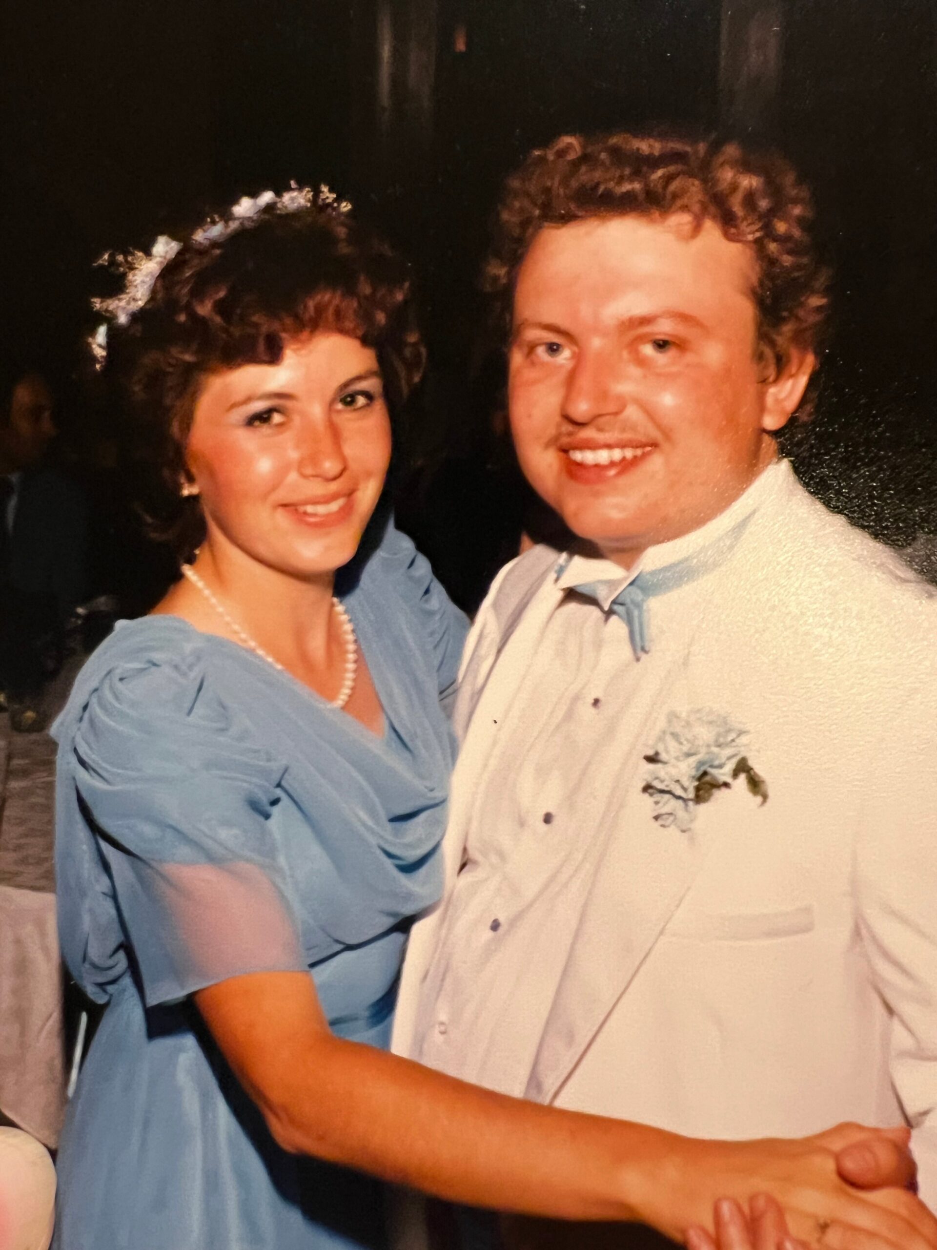 Smiling couple dressed for a formal event pose together in a dimly lit setting, arm in arm.