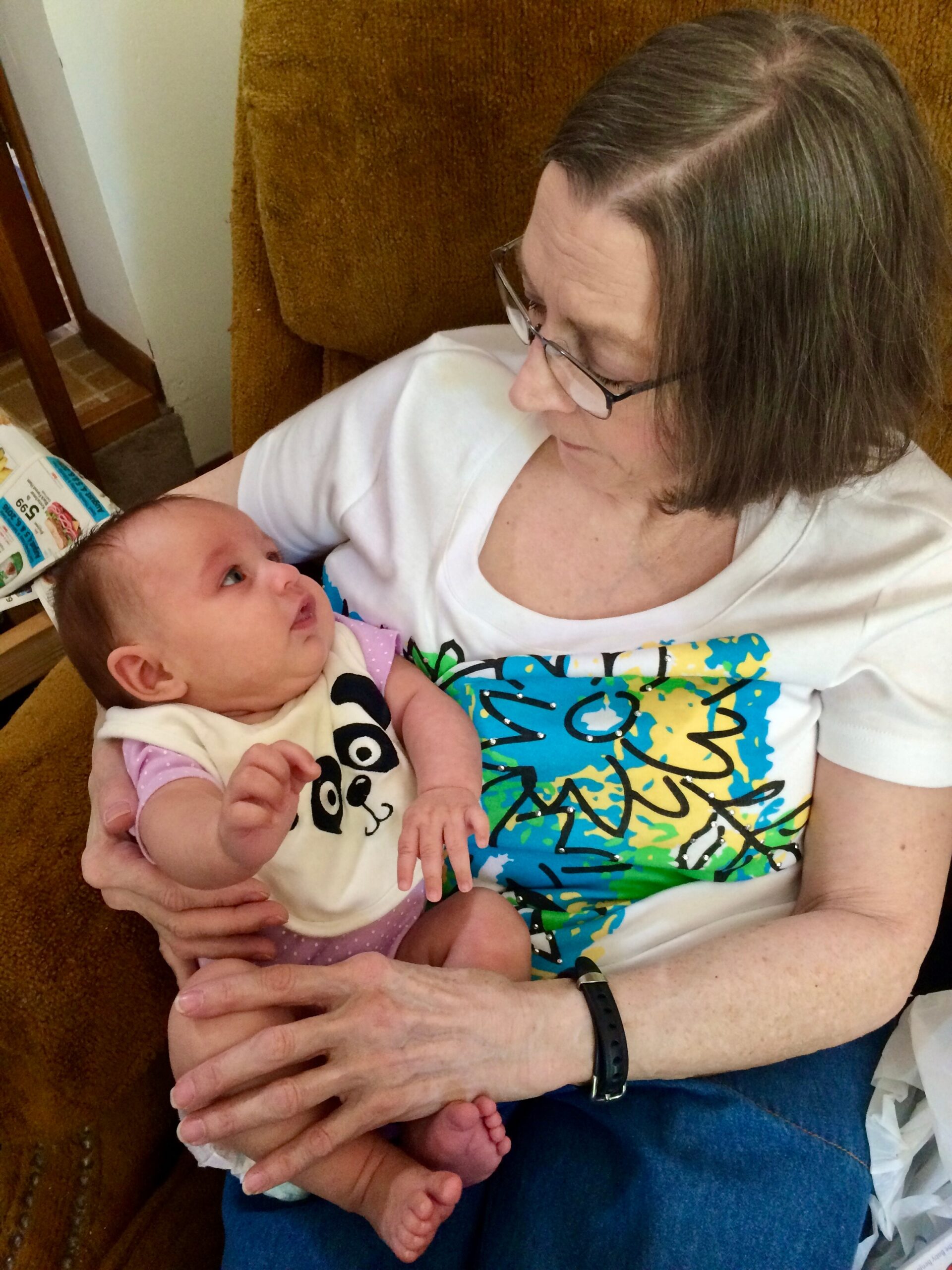 Grandmother with glasses sits on a brown couch, cradling a newborn baby in a pale outfit with a panda face.