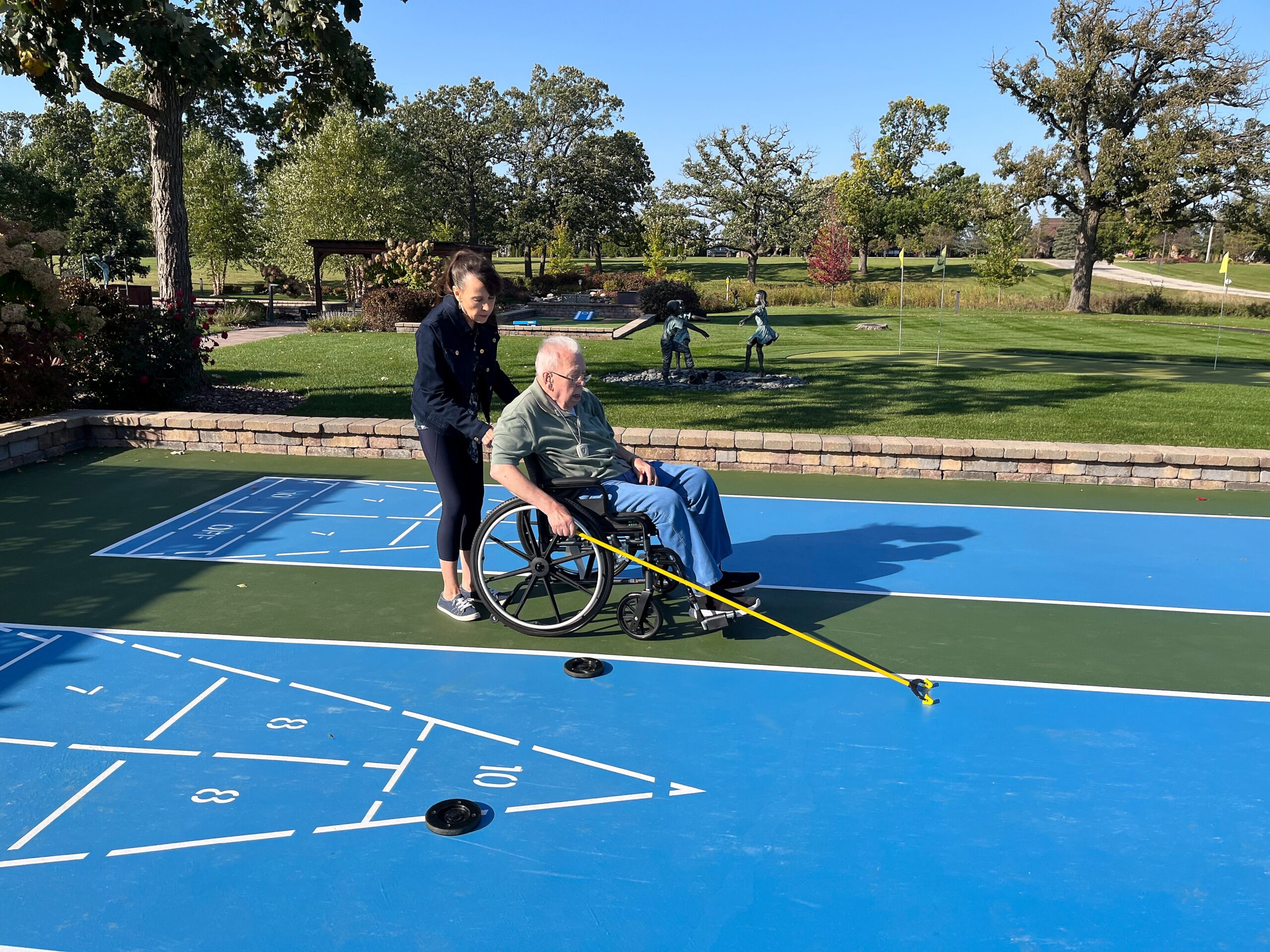 Caregiver assists an elderly man in a wheelchair on a bright blue outdoor game court with numbered lanes or markings, in a park setting.
