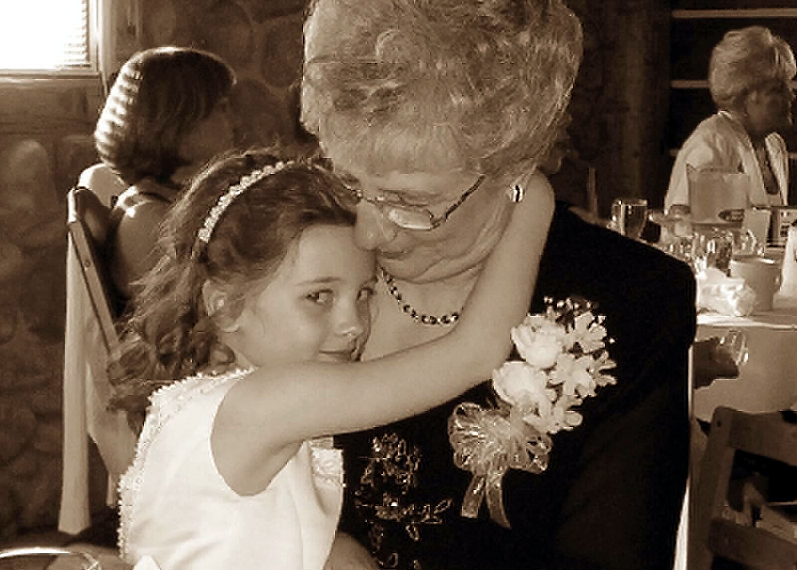 Young girl in a white dress hugs an older woman at a wedding reception, both smiling.