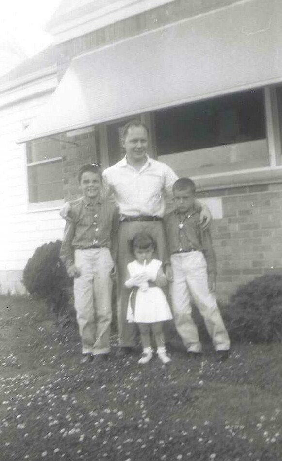 Man with two boys and a girl standing outdoors in front of a brick house, with the man’s arms around the children.