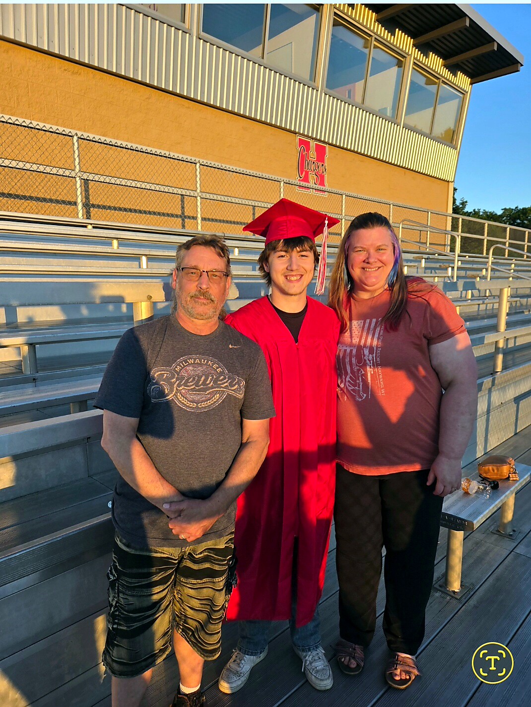 Graduate in a red cap and gown stands between a man in a dark t-shirt and a woman in a pink shirt on metal bleachers outside a stadium at sunset.