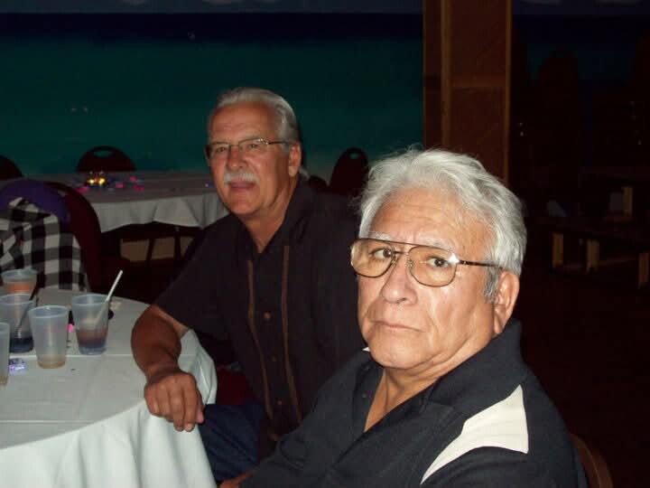 Two older men sit at a table with cups and straws in a dim dining area, posing for the camera.