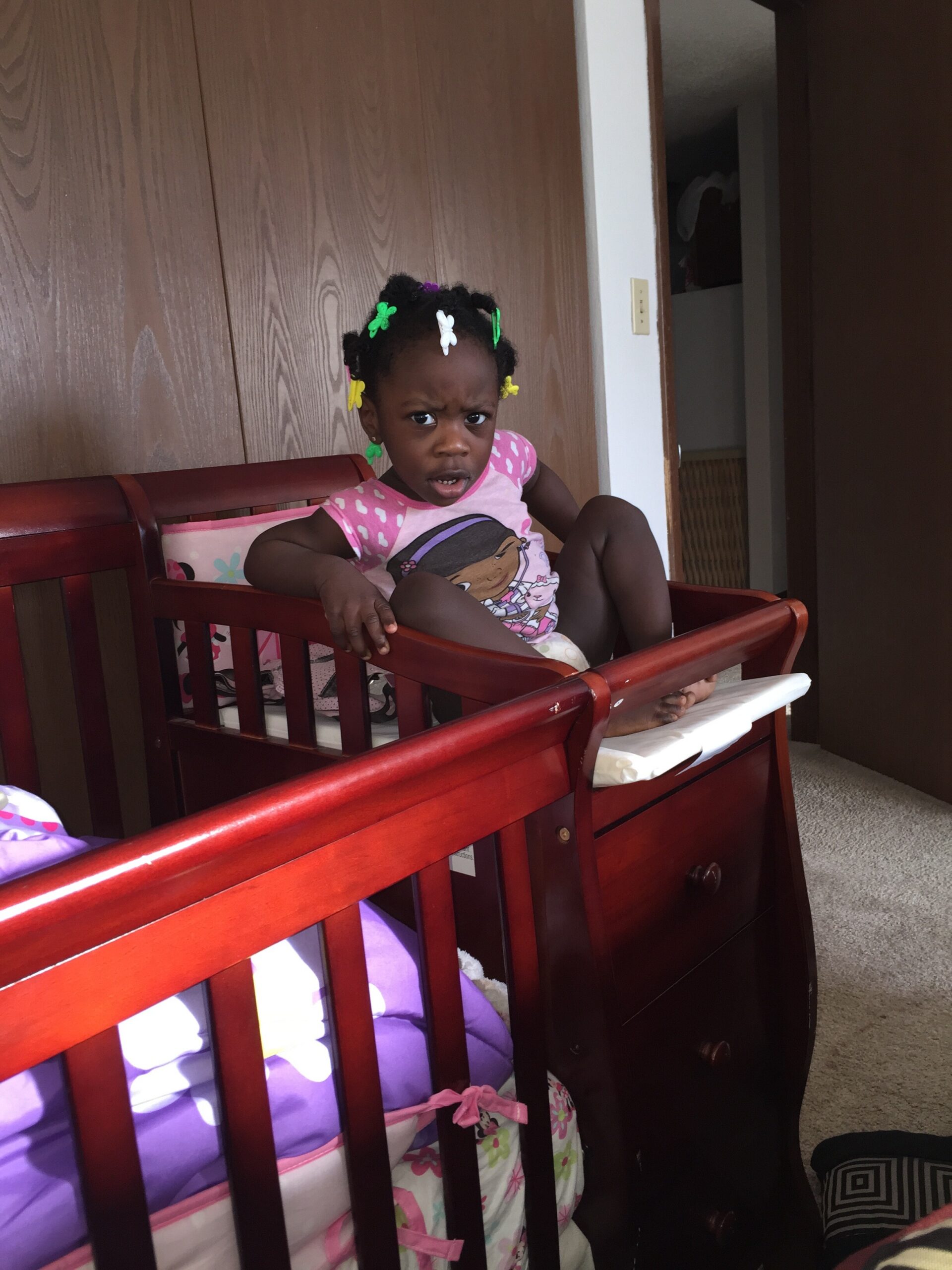 Young girl in a pink shirt sits in a red wooden crib, frowning with colorful hair clips in her braids in a bedroom.