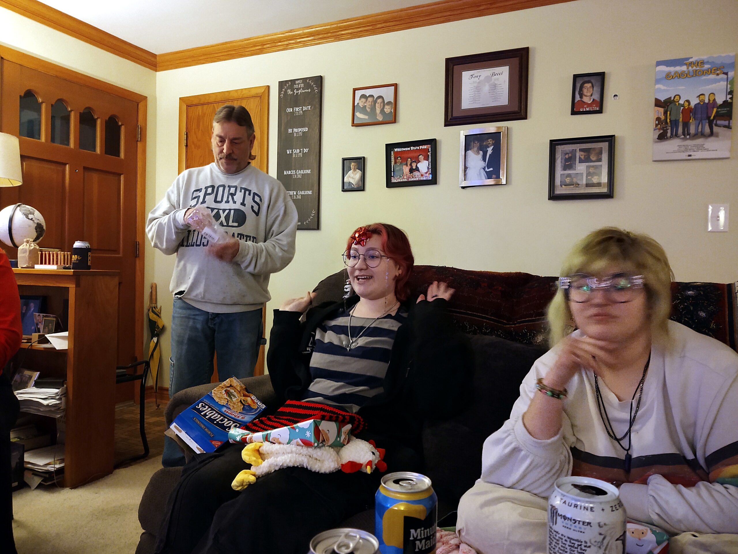 Man standing with a bottle in a living room during a party, others seated, gifts and cans on the table behind him.