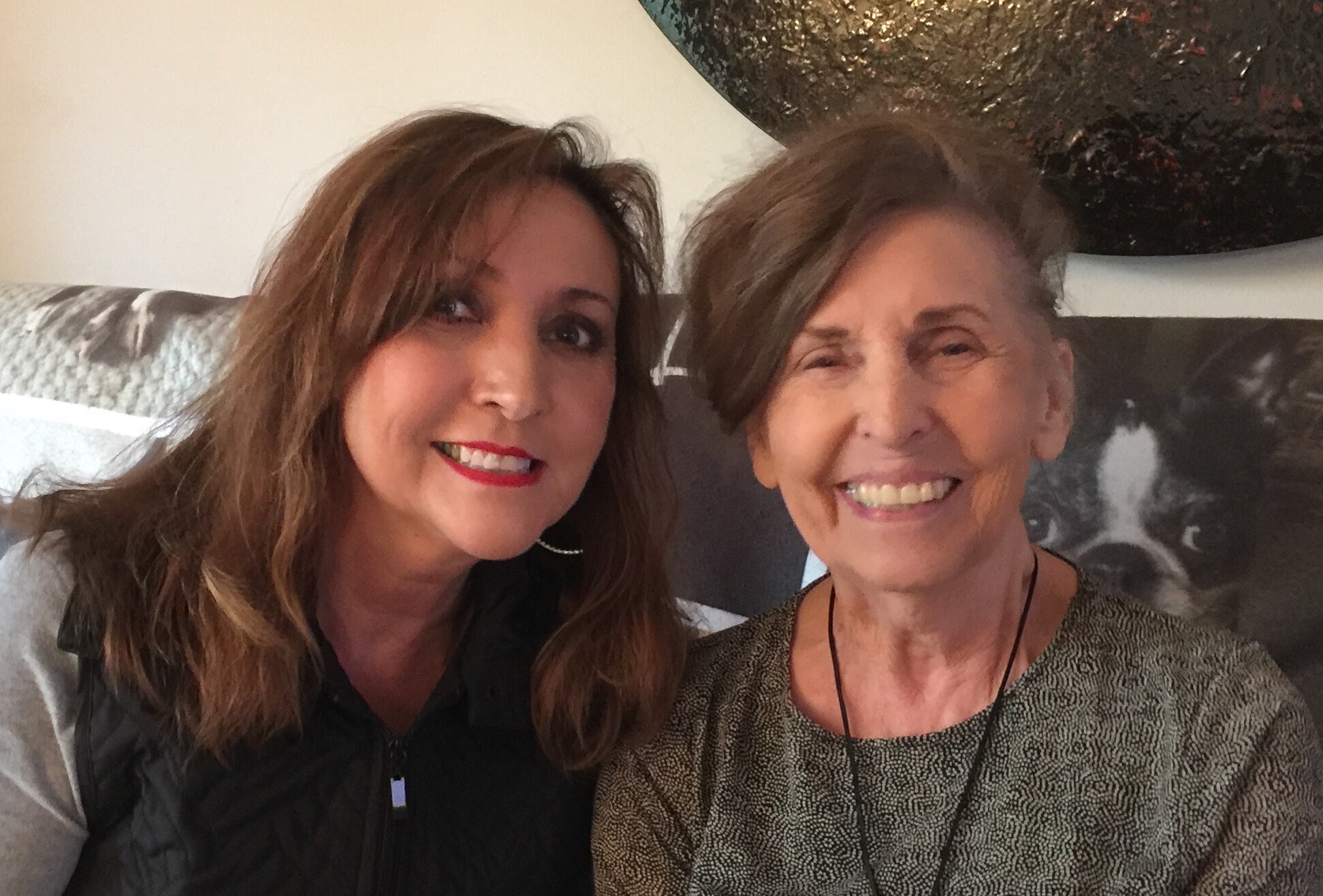 Two women smiling for a selfie on a sofa, with abstract art in the background on the wall behind them.