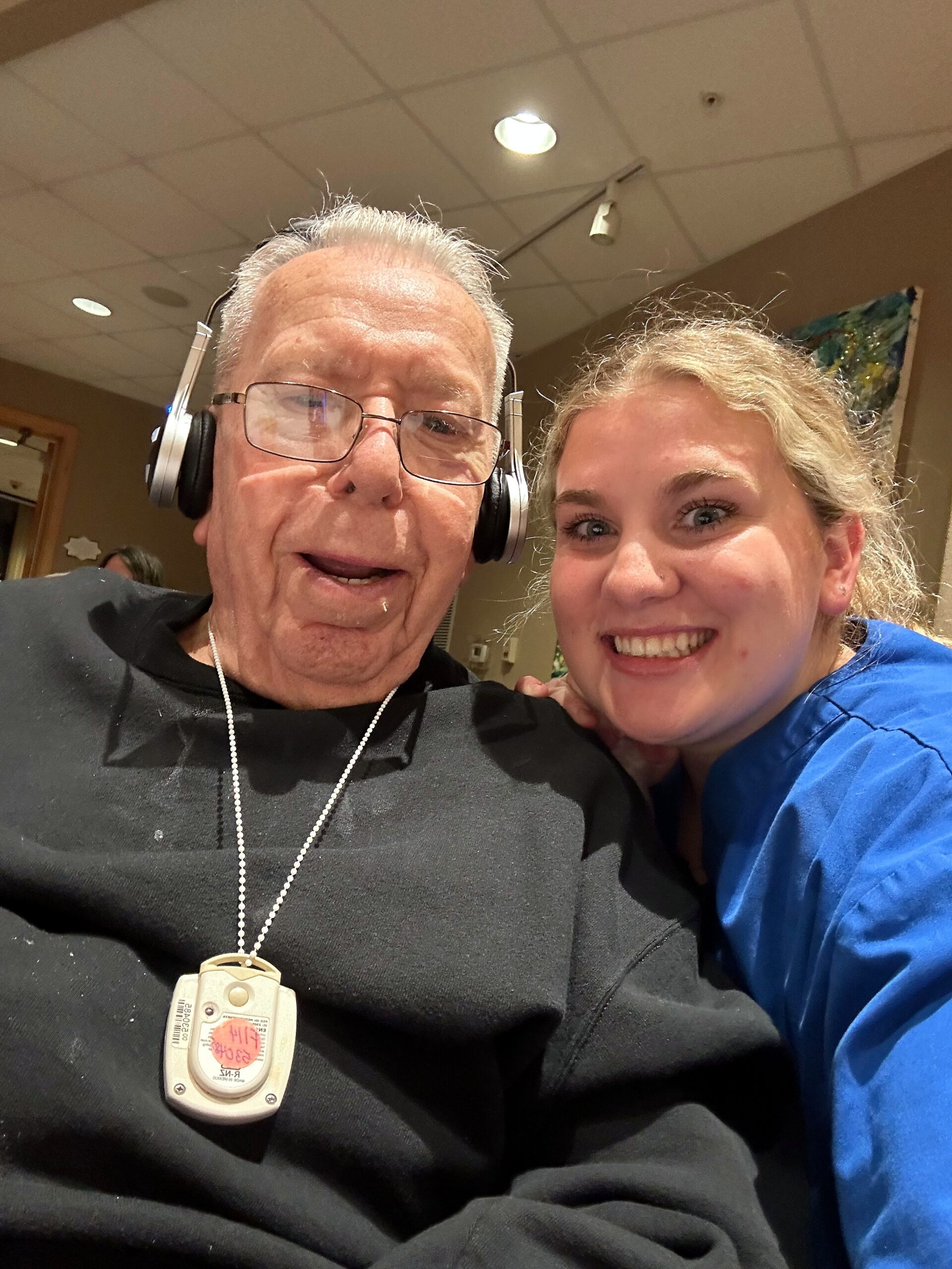Smiling older man with over-ear headphones and a medical device on a lanyard, beside a young woman in blue scrubs.