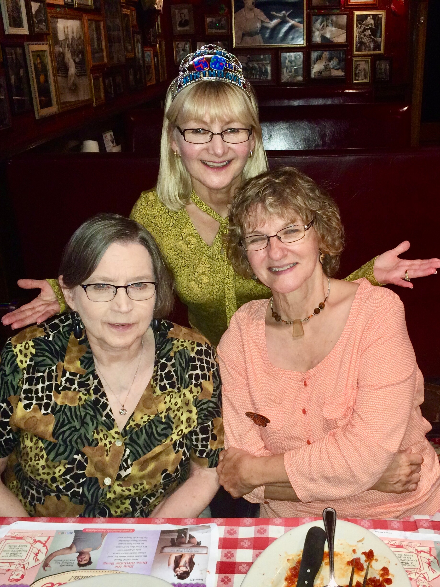Three women posing at a restaurant table; center woman wears a jeweled crown and a green patterned top.