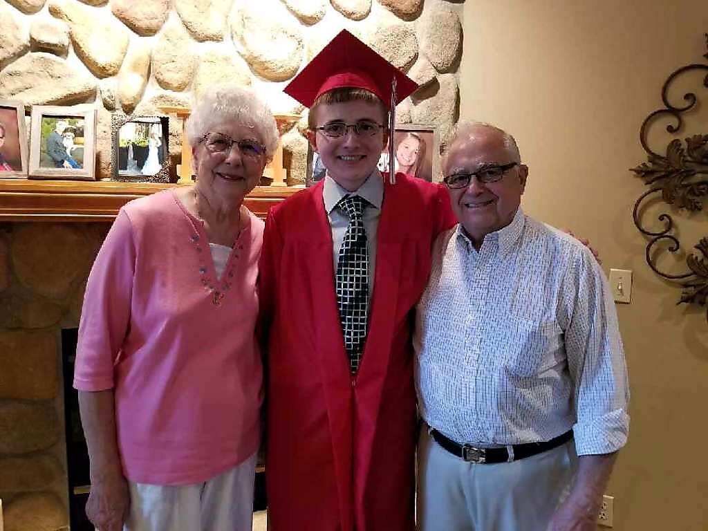 Graduate in a red cap and gown stands between an elderly woman in pink and an elderly man in a striped shirt, posing in a living room by a stone fireplace.