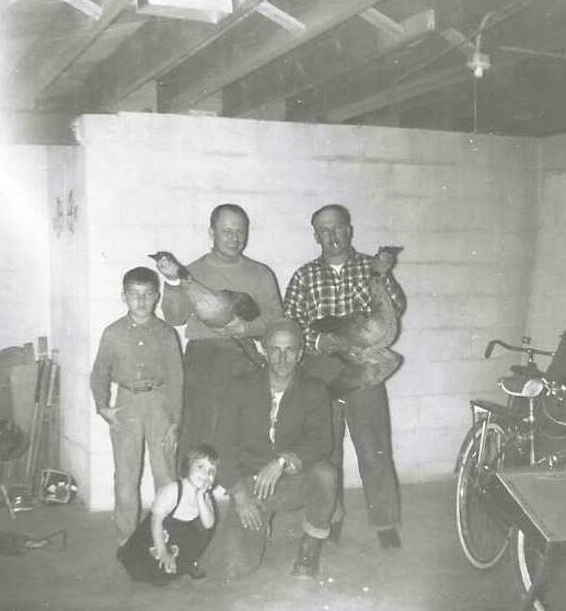 Group portrait in a garage: two men hold small dogs, a kneeling child, a man crouches, another man stands; a bicycle is on the right.