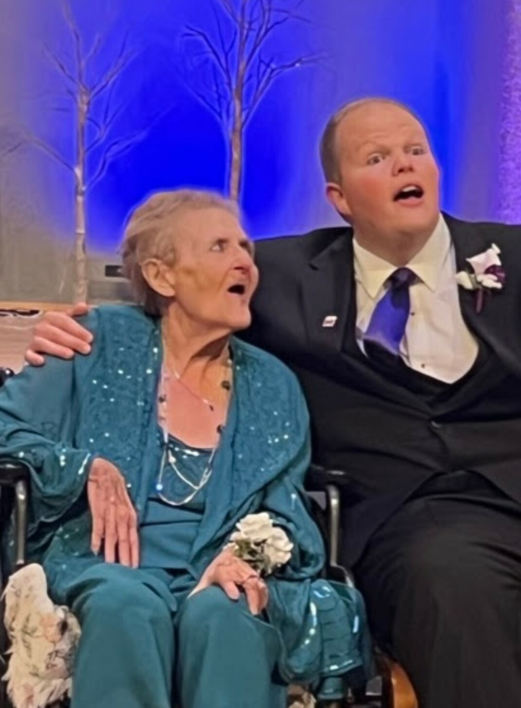 Elderly woman in a teal sequined gown sitting in a wheelchair, with a man in a suit beside her, both looking surprised on stage with a blue backdrop.