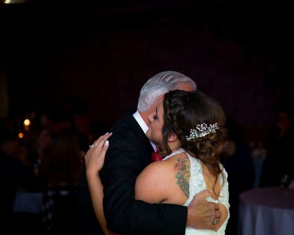 Bride and groom embrace and kiss during a wedding reception dance; bride in a white lace dress with a jeweled hair accessory, tattoo visible on her back/shoulder area in the dim venue lighting.
