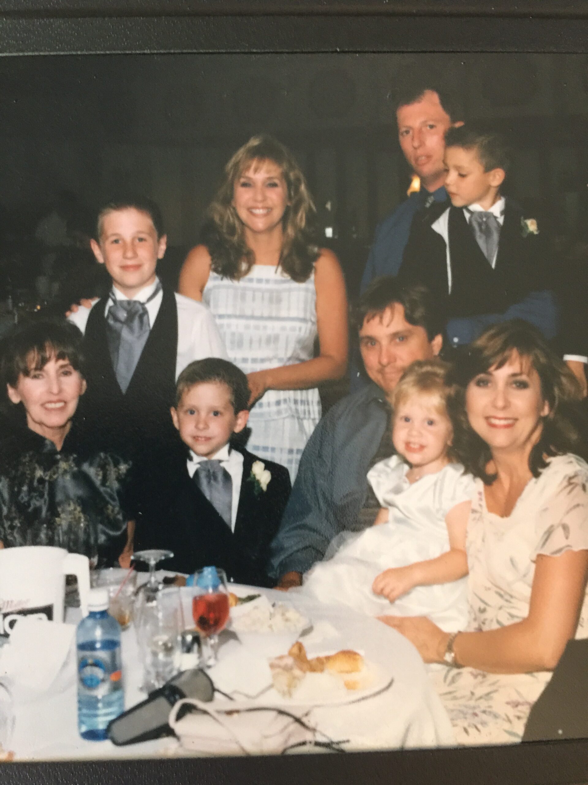 Family group photo at a formal dinner, with a smiling woman in a white dress, kids in suits, and adults around a table with beverages and food подпис.