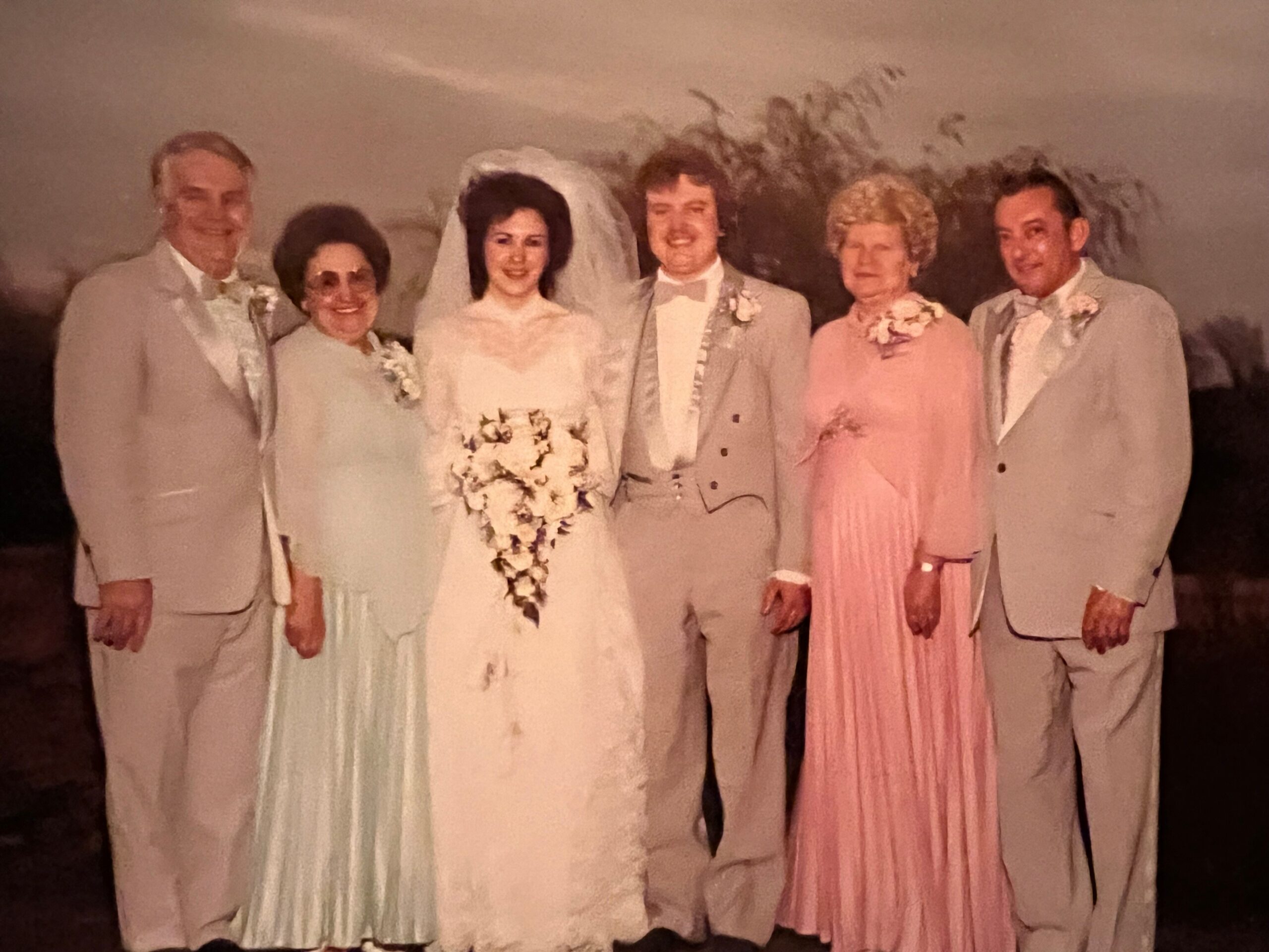 Bride in white gown holding a cascading bouquet stands beside groom in a light suit; surrounded by four guests in pastel formal wear, posing for a group wedding photo.